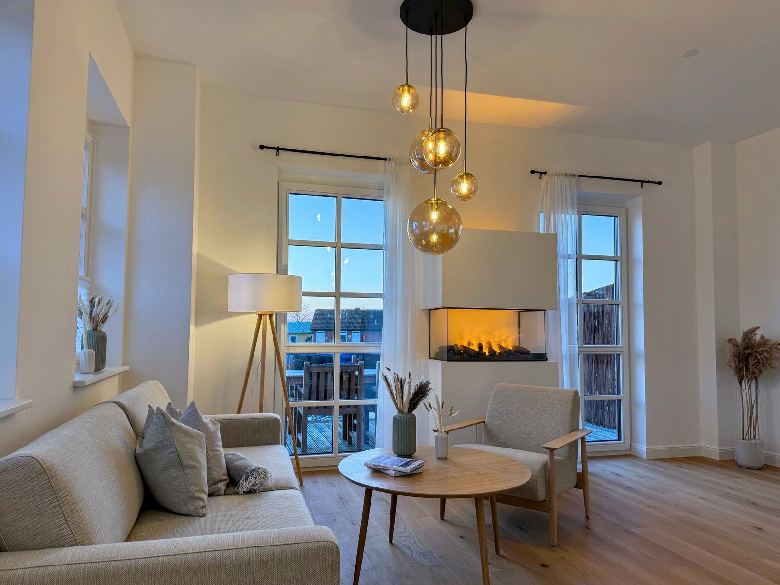 Wohnzimmer mit Sofa, Sessel, Tisch und Kamin. Fenster mit Blick auf Terrasse.