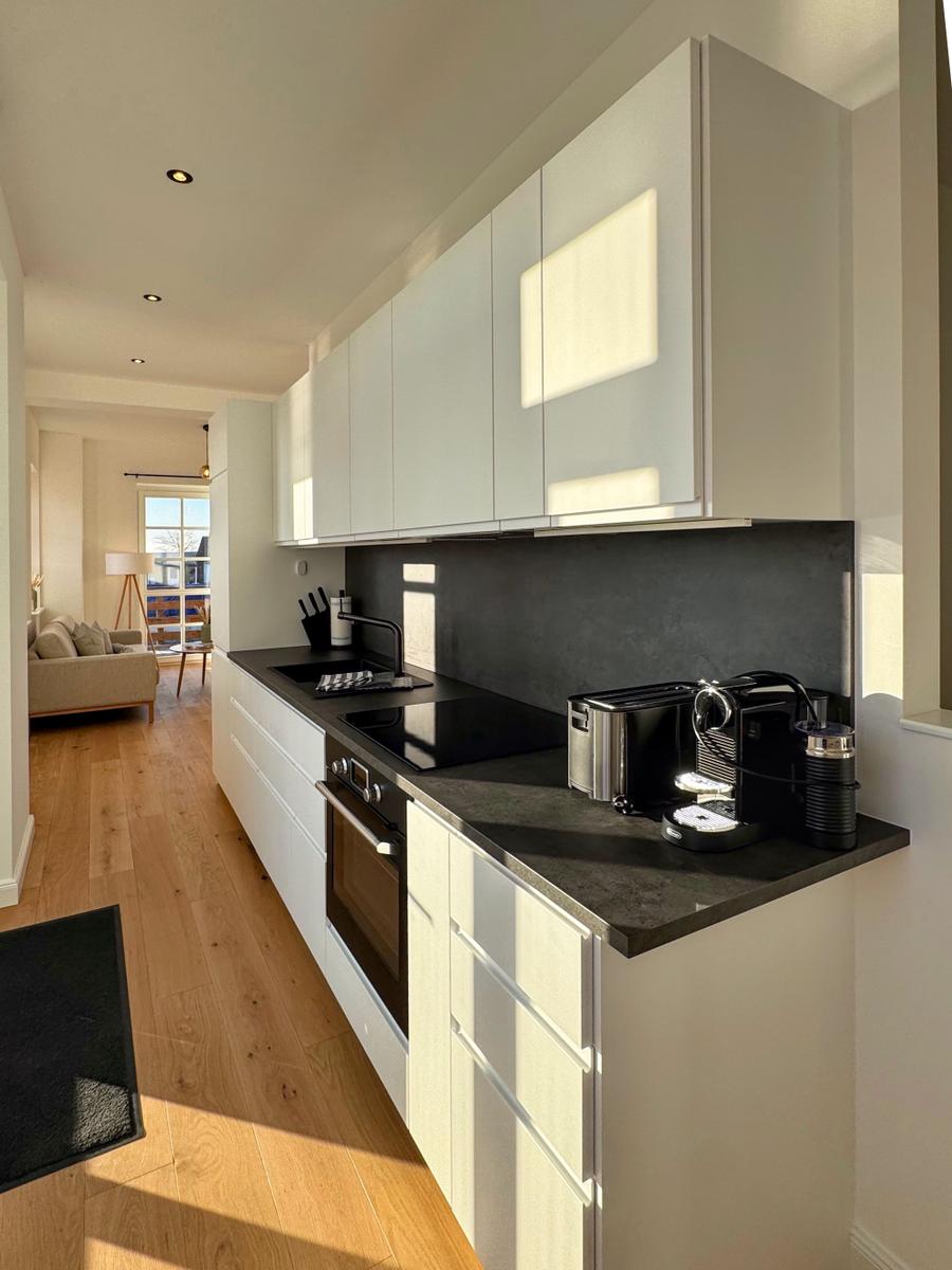 Moderne Küche mit weißen Schränken und dunkler Arbeitsplatte. Blick in das Wohnzimmer.