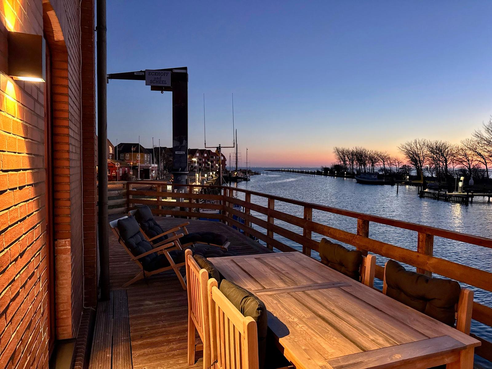 Terrasse mit Tisch und Stühlen an Wasser, Blick auf Hafen und Sonnenuntergang.
