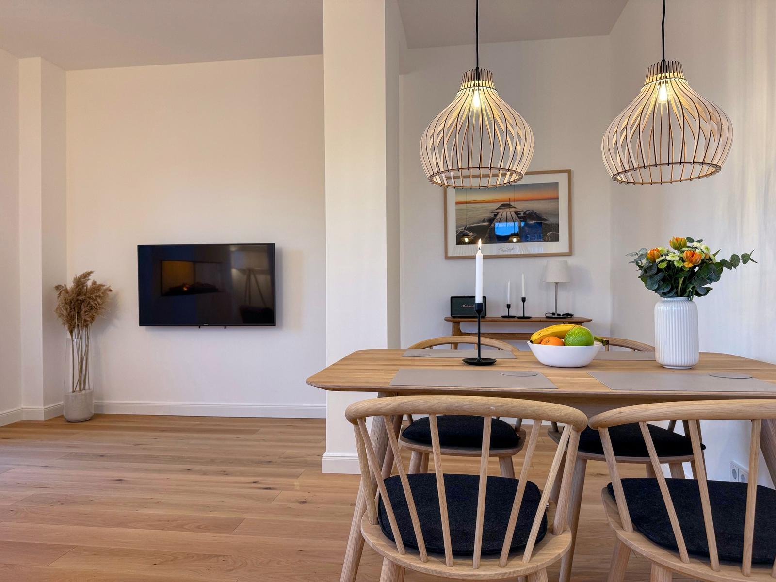 Esszimmer mit Holztisch, Stühlen und zwei Hängelampen. Fernseher an der Wand.