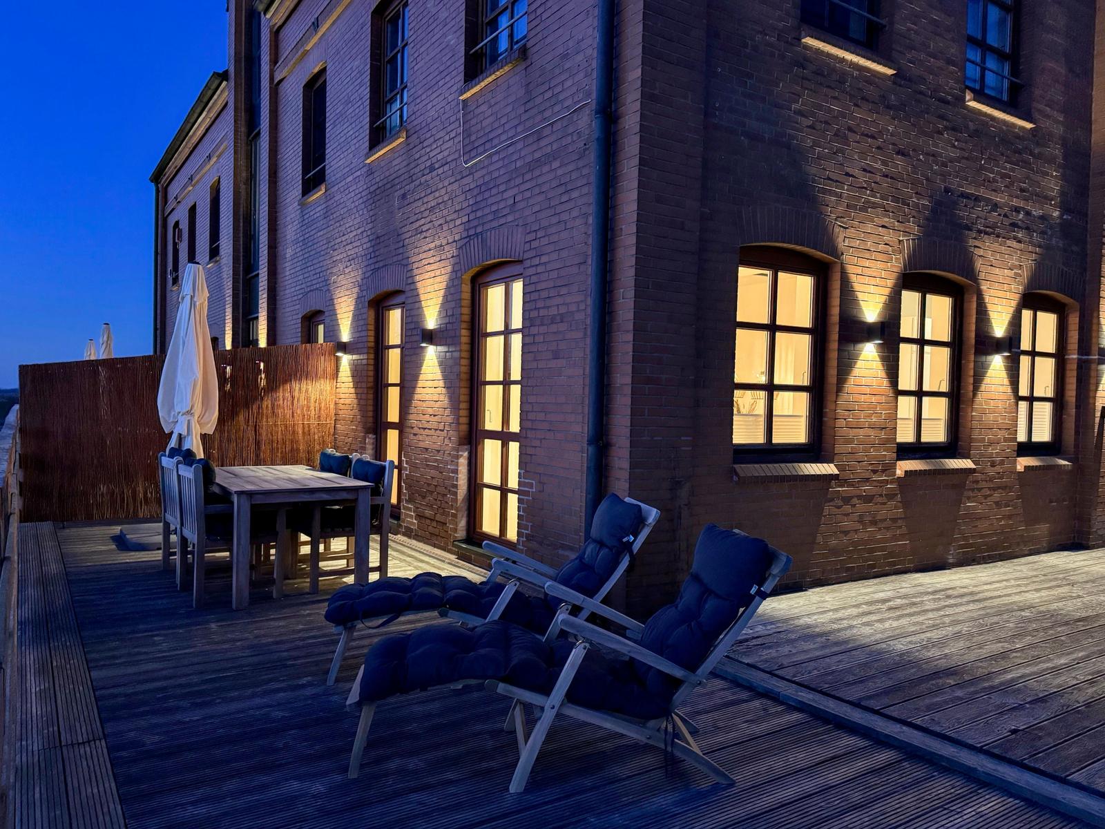 Terrasse mit Liegestühlen und Tisch vor Backsteinhaus bei Dämmerung.