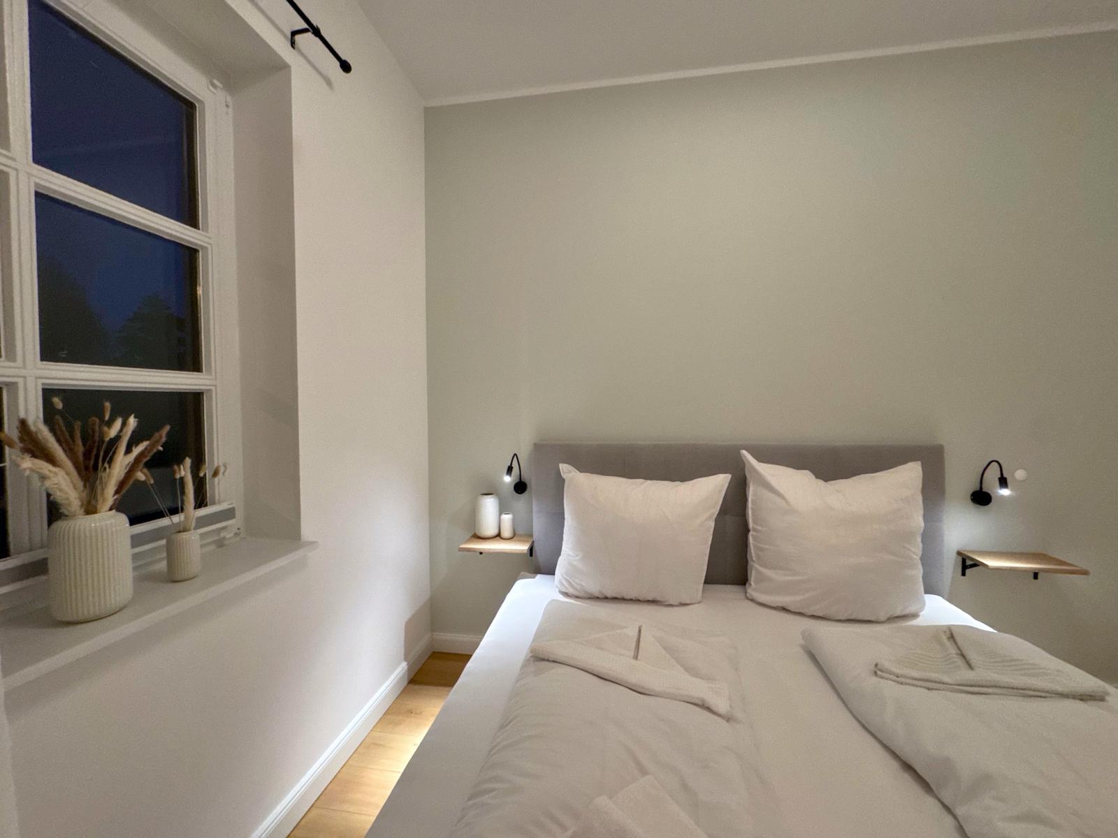 Schlafzimmer mit Bett, zwei Nachttischen und Fenster mit Pflanzen.