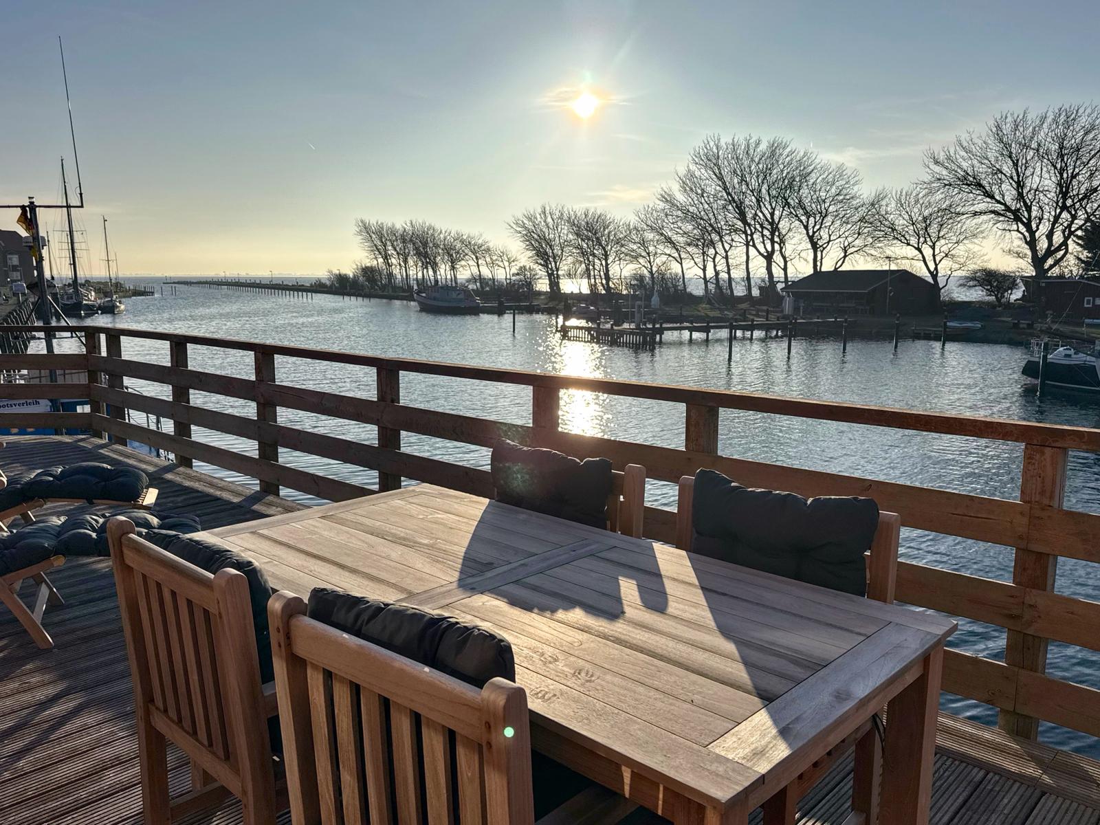 Holztisch mit Stühlen auf Terrasse mit Blick auf Wasser und Boote.