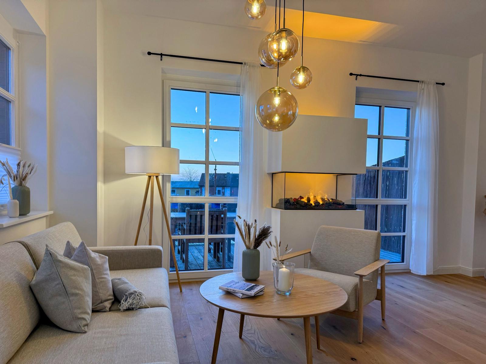 Wohnzimmer mit Sofa, Sessel, Kamin und Holzboden. Fenster und Hängelampe sichtbar.