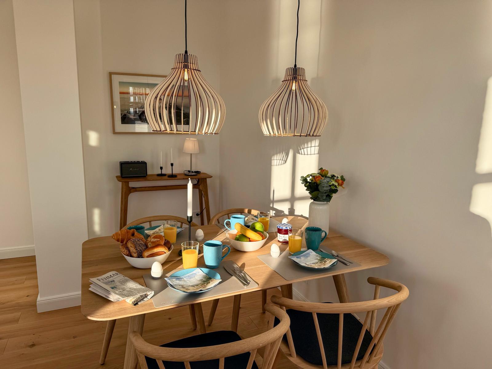 Esszimmer mit Holztisch, Stühlen und hängenden Lampen. Tisch gedeckt mit Frühstück.