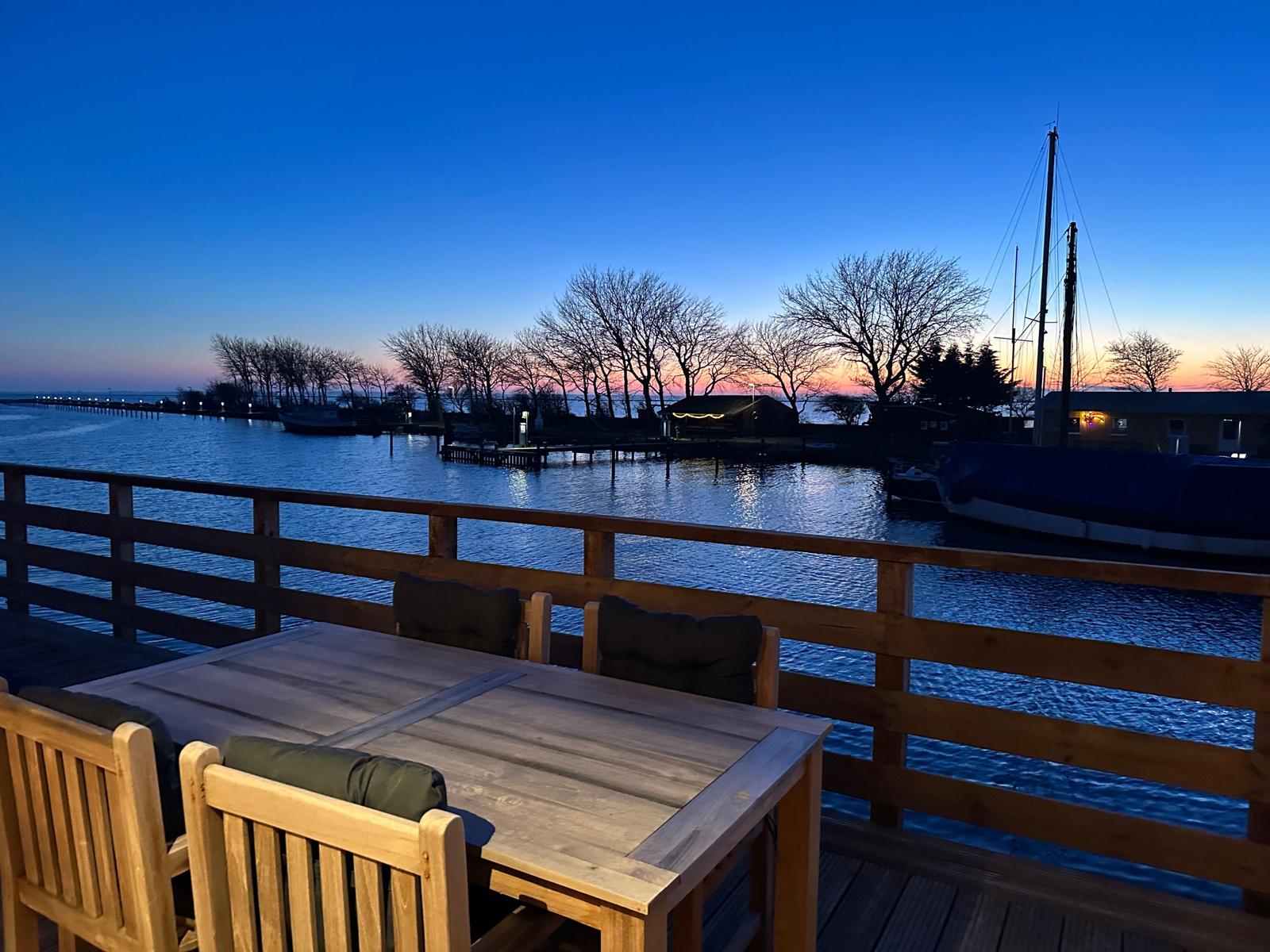 Terrasse mit Tisch und Stühlen an Wasserfront bei Sonnenuntergang.