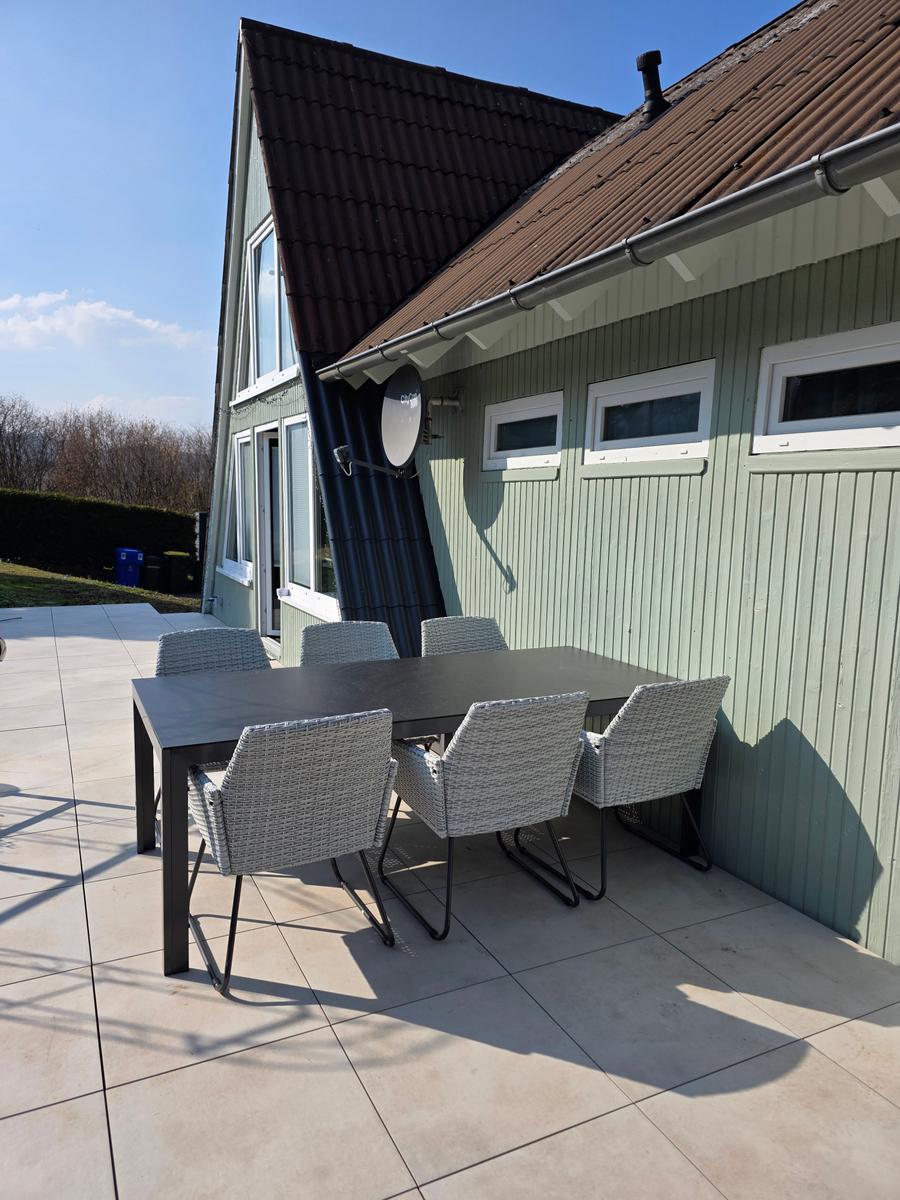 Terrasse mit Tisch und sechs Stühlen vor einem Haus mit grünem Holzverkleidung.