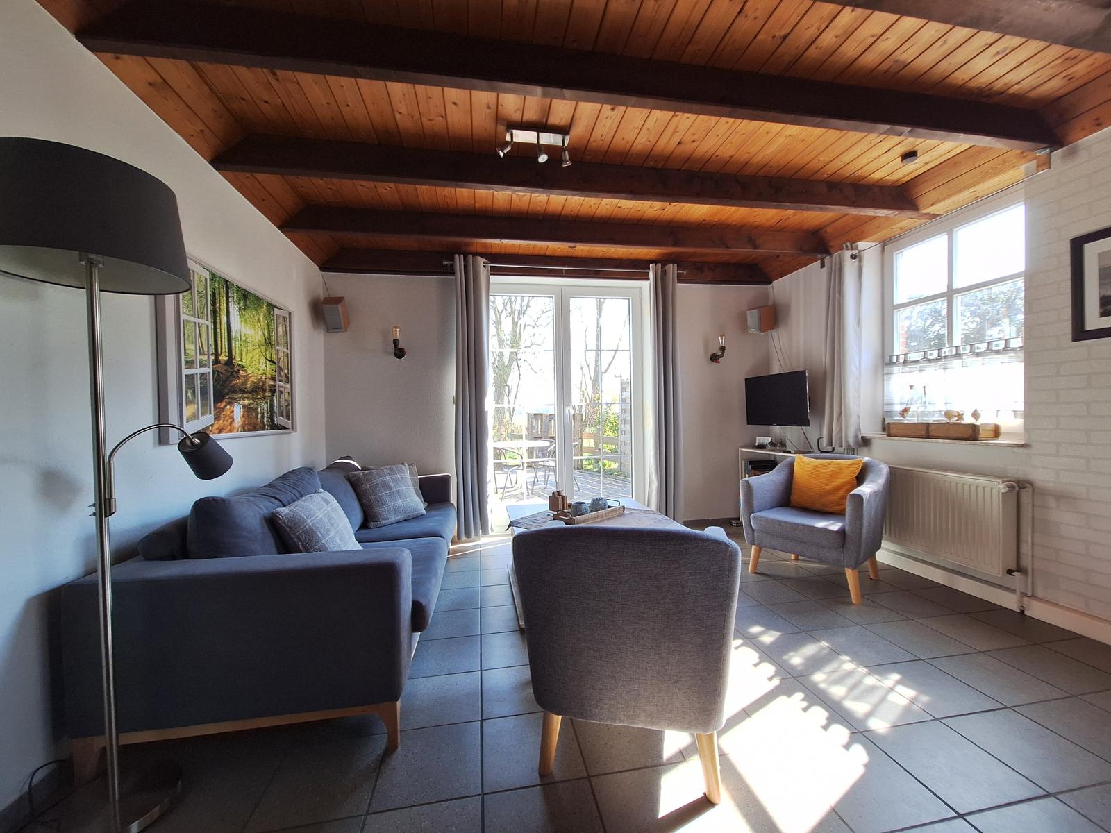 Wohnzimmer mit Sofa, Sessel und TV. Holzdecke und große Fenster.