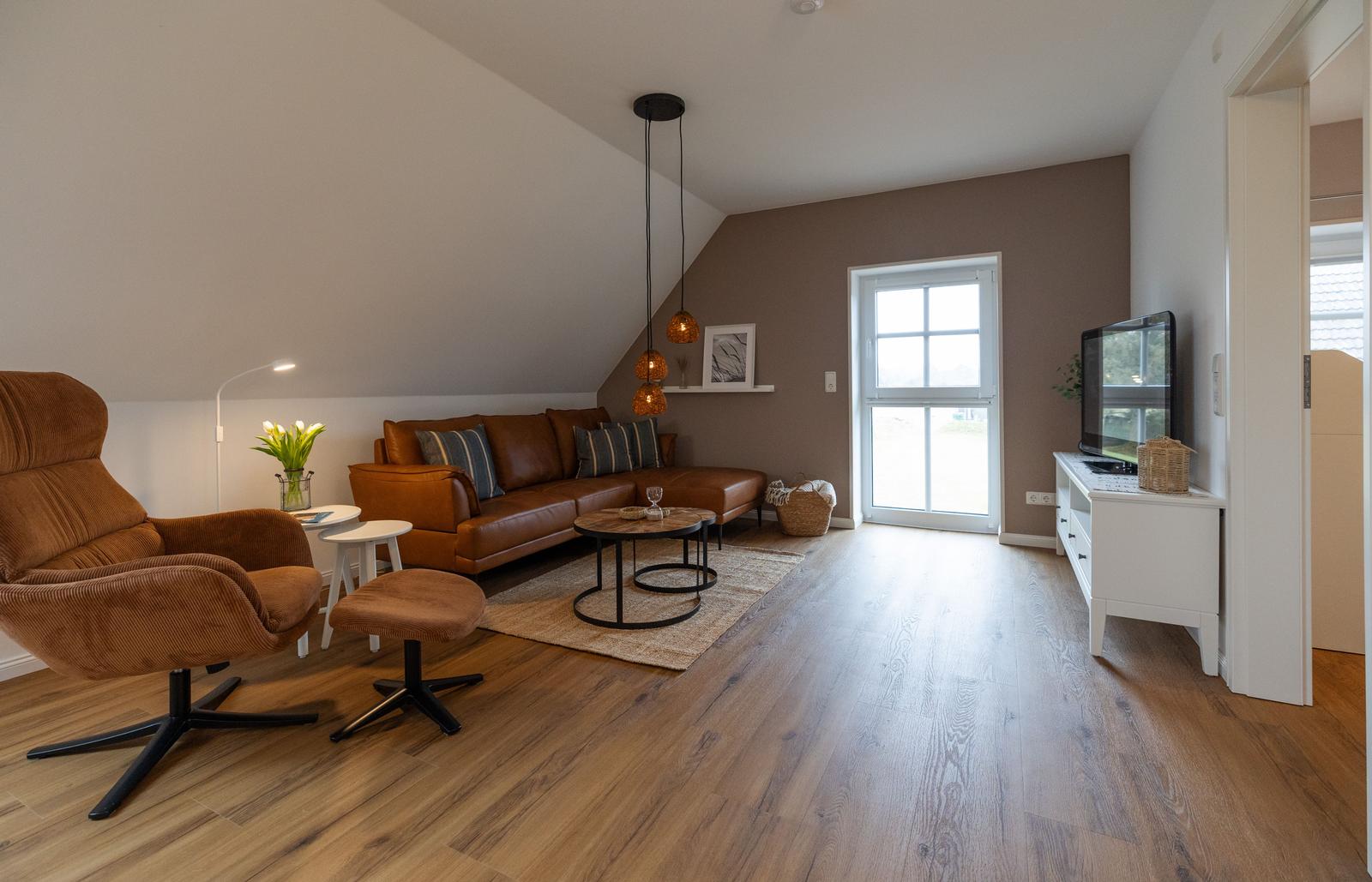 Wohnzimmer mit braunem Sofa, Sessel und Holzboden. Fenster und TV-Schrank sichtbar.