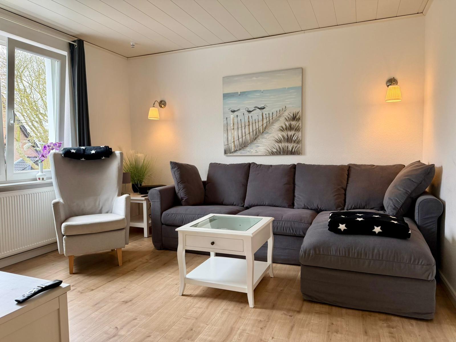 Ein Wohnzimmer mit grasem Sofa, weißem Couchtisch und Wandbild. Fenster mit Blick nach draußen.