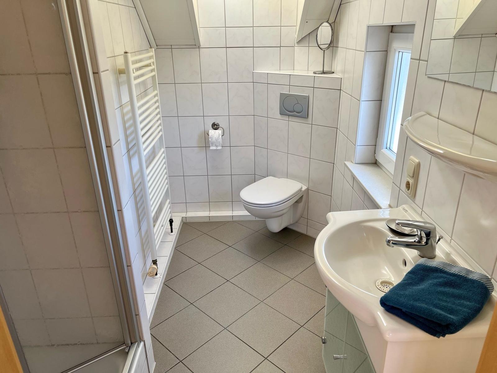 Badezimmer mit Toilette, Waschbecken und Dusche. Weiße Fliesen und blauer Handtuch.