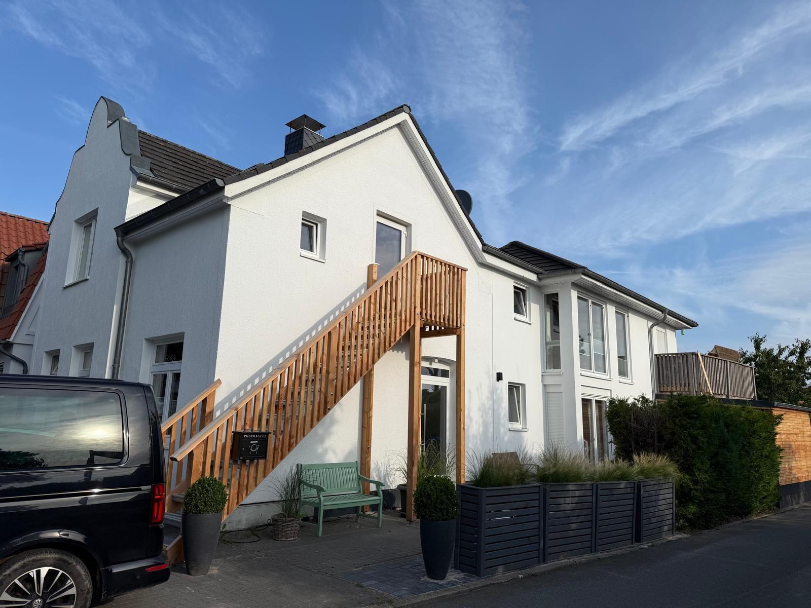 Weißes Haus mit Holztreppe und Balkon. Grünfarbene Bank vor Eingang.