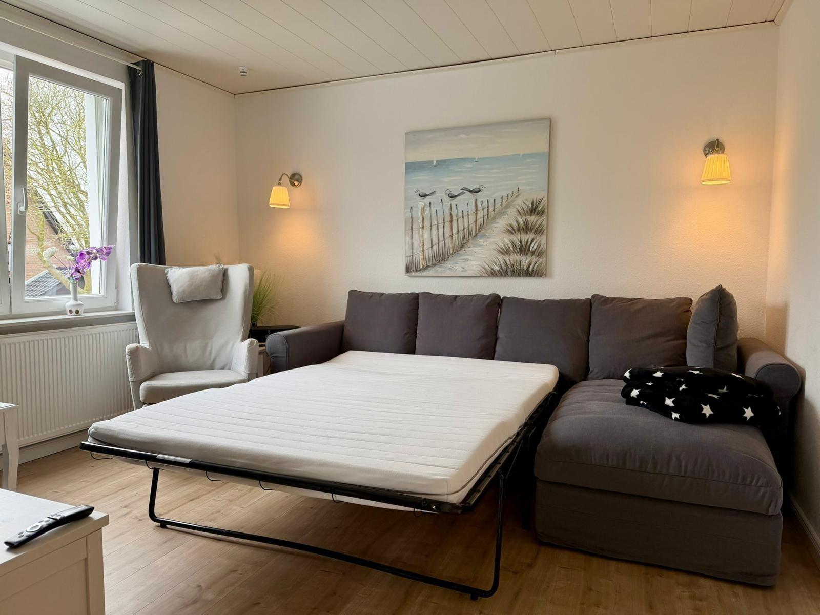 Wohnzimmer mit Sofa, Schlafmatratze und Wandbild. Fenster mit Blick nach draußen.