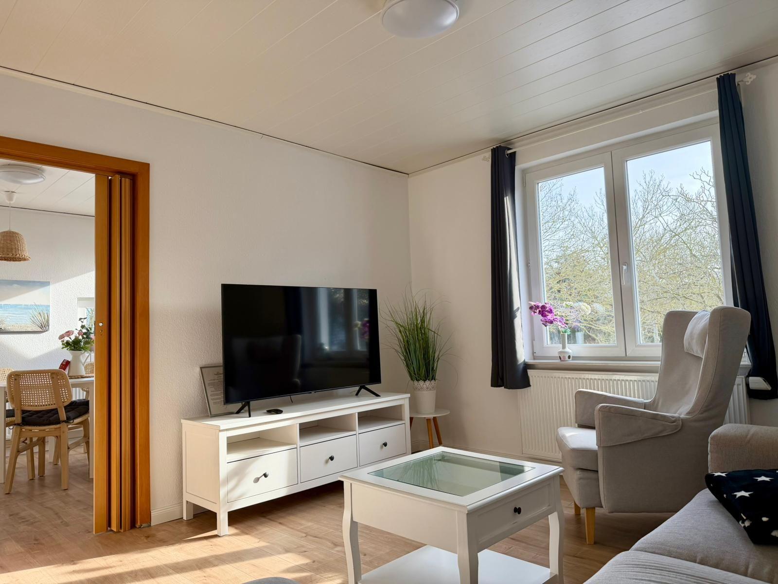 Wohnzimmer mit Fernseher, Sessel und Tisch. Durchgang ins Esszimmer. Fenster mit Blick auf Bäume.