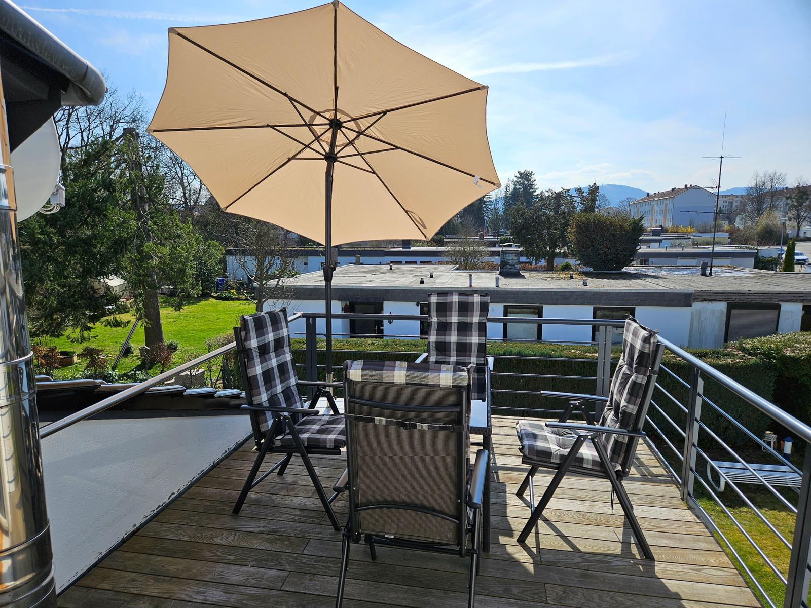 Terrasse mit Sonnenschirm und vier Stühlen. Blick auf Grünfläche und Gebäude.