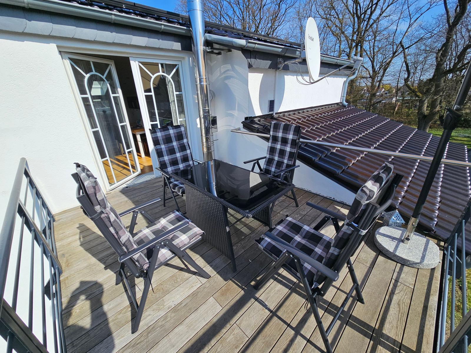 Terrasse mit vier Stühlen und Tisch. Hintergrund: Haus mit Satellitenschüssel und Dach.