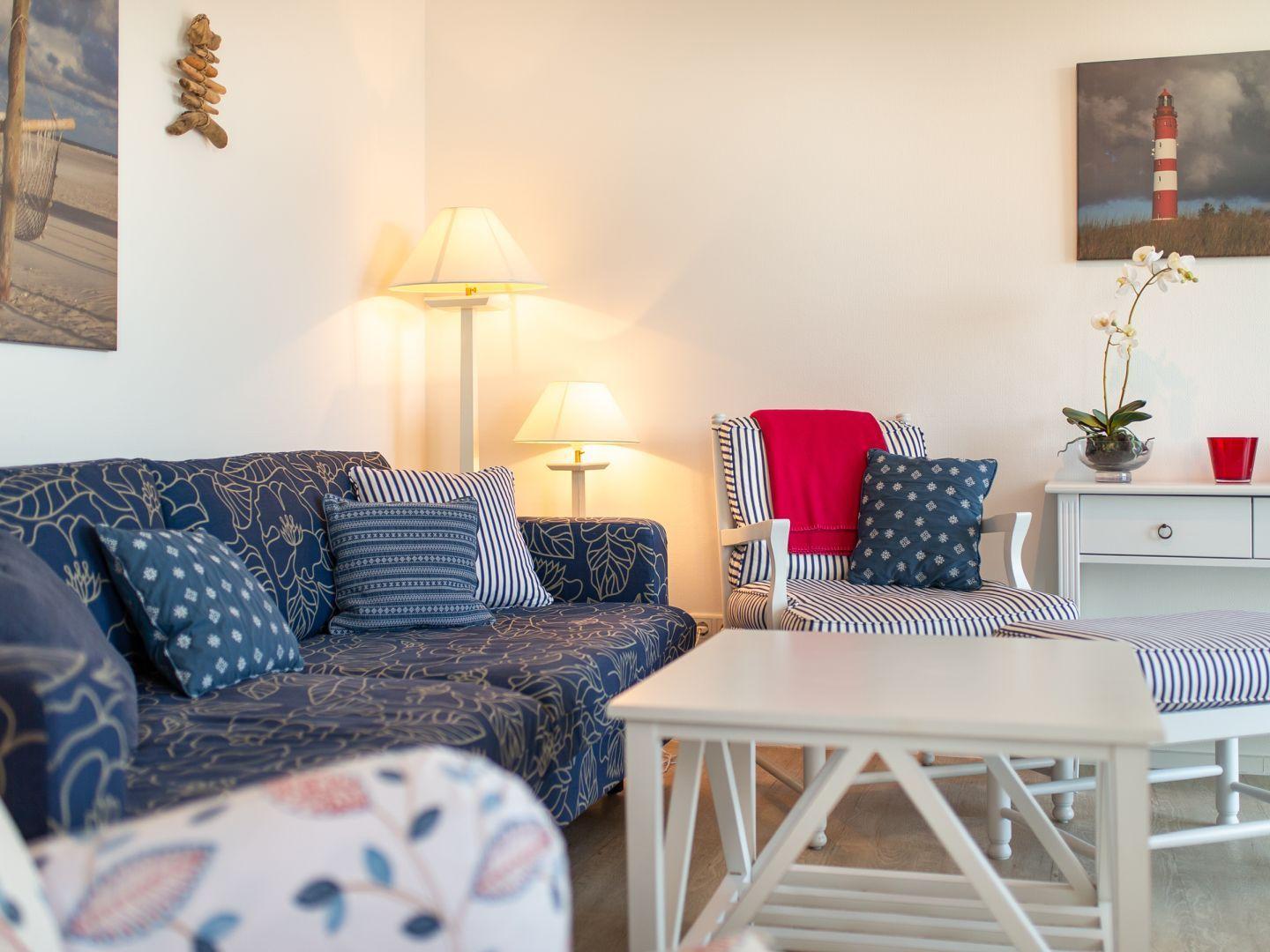Wohnzimmer mit Sofa, Sessel und Tisch. Wandbilder zeigen Leuchtturm und Strand.