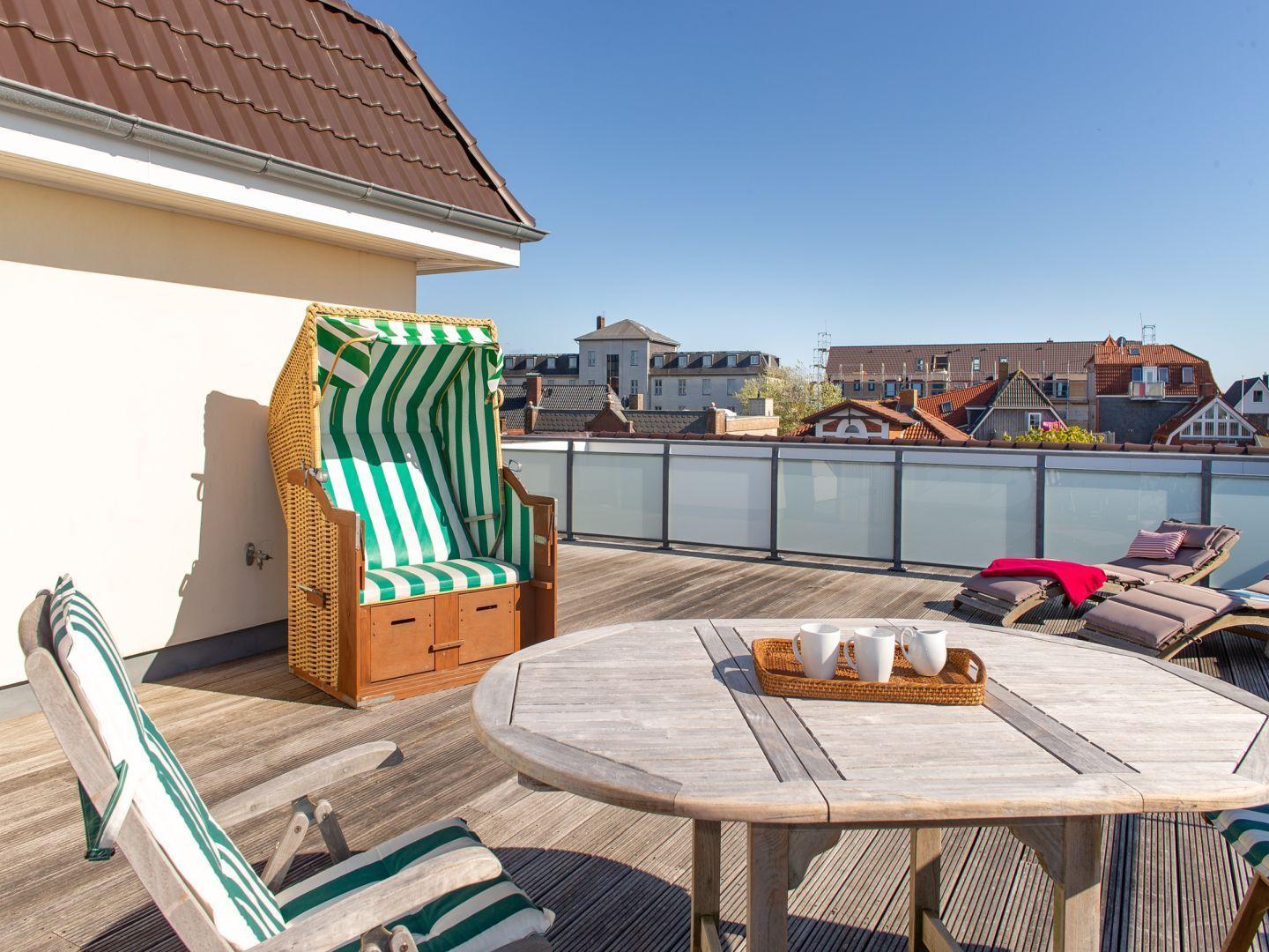 Dachterrasse mit Strandkorb, Tisch und Liegestühlen. Blick auf Stadt und Dächer.