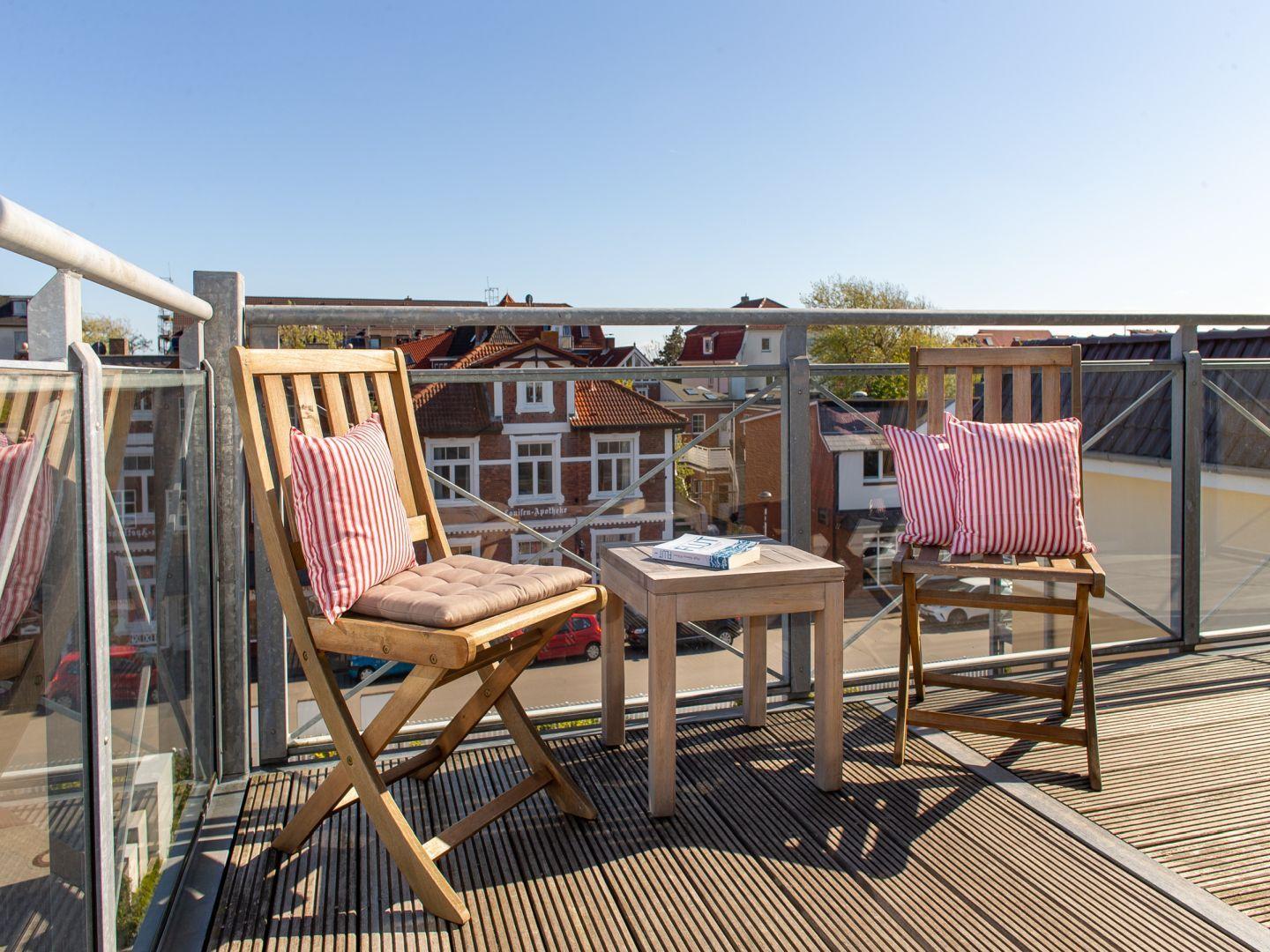Terrasse mit zwei Holzstühlen, Tisch und roten Kissen. Blick auf Wohnhäuser und Straße.