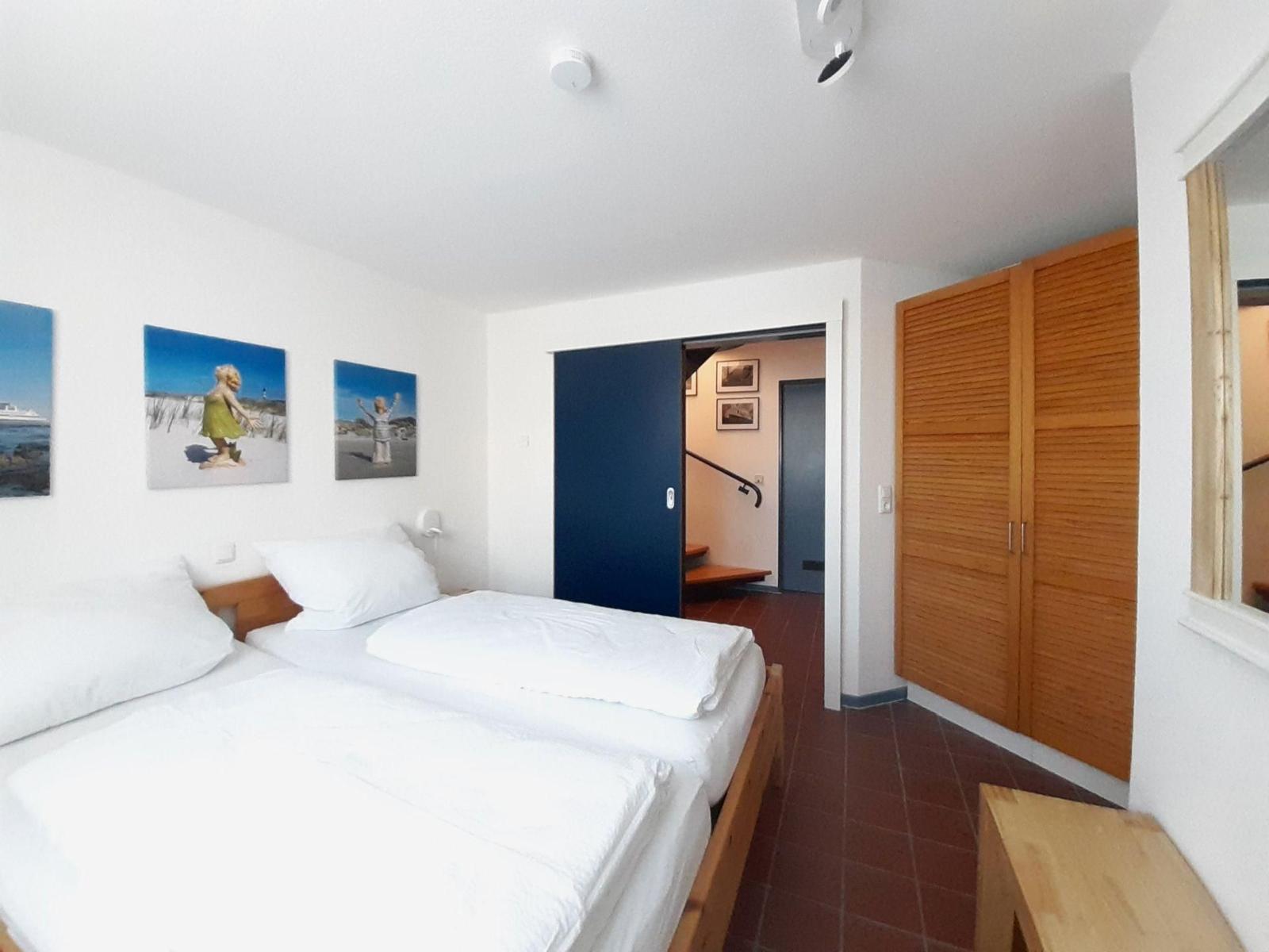 Zimmer mit Doppelbett, Holzschrank und Wandbildern. Tür führt in einen Flur mit Treppe.
