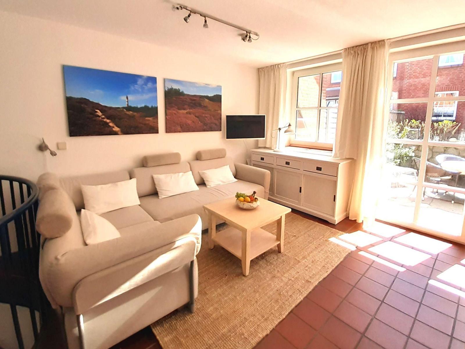 Wohnzimmer mit Sofa, Couchtisch und TV. Fenster und Glastür führen auf eine Terrasse.