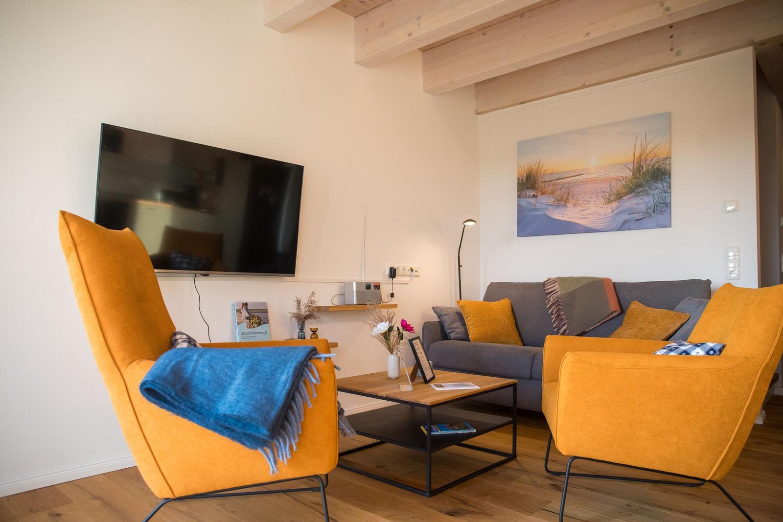 Wohnzimmer mit grauem Sofa, zwei orangefarbenen Sesseln und Fernseher an der Wand.