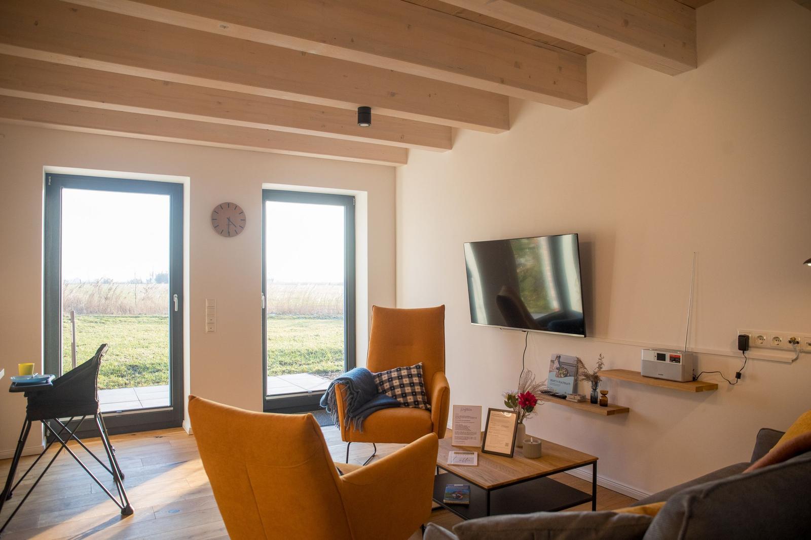 Wohnzimmer mit Holzbalken, TV, zwei orangefarbenen Sesseln und Blick auf Grünfläche.