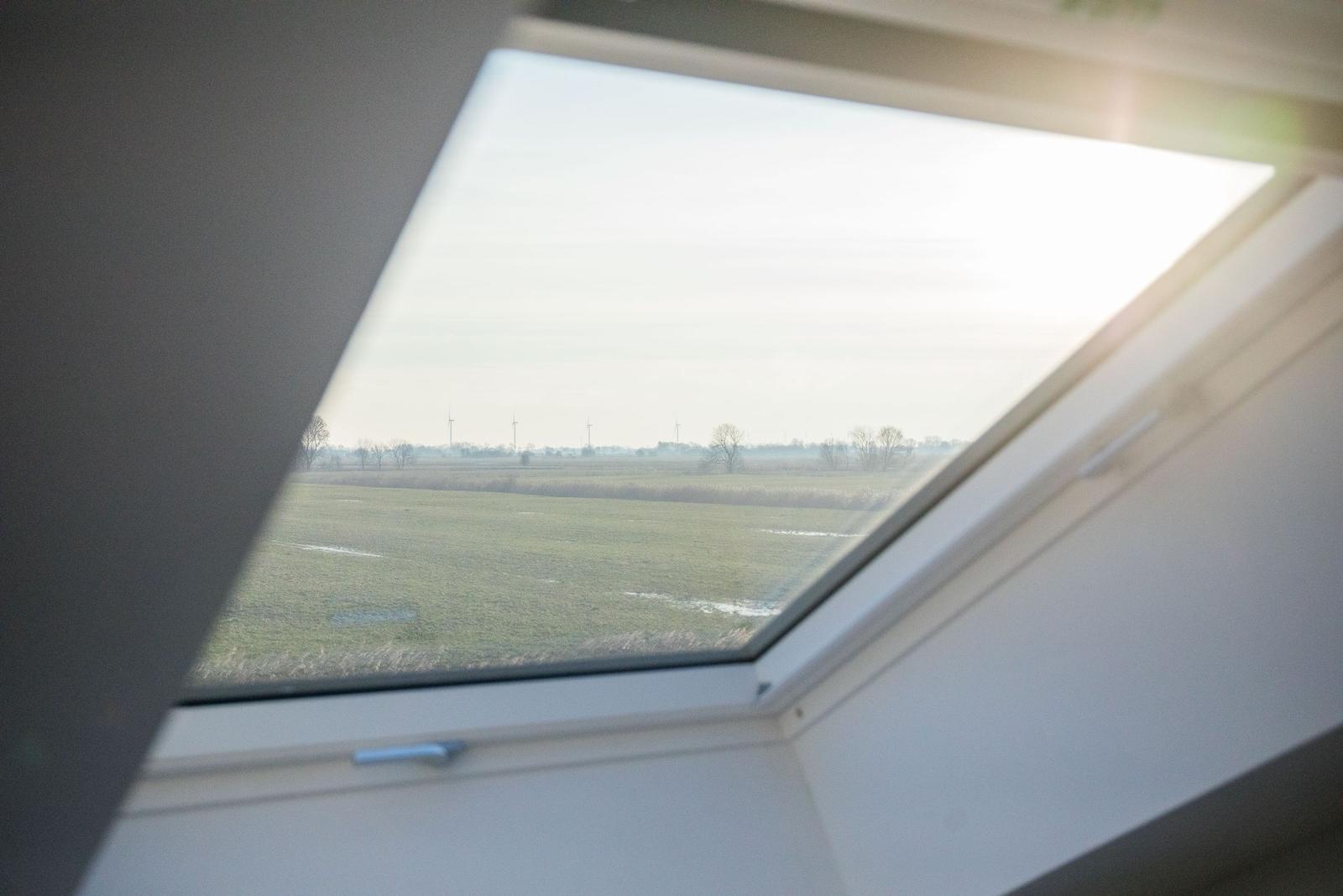 Dachfenster mit Blick auf grünes Feld und Windräder im Hintergrund.