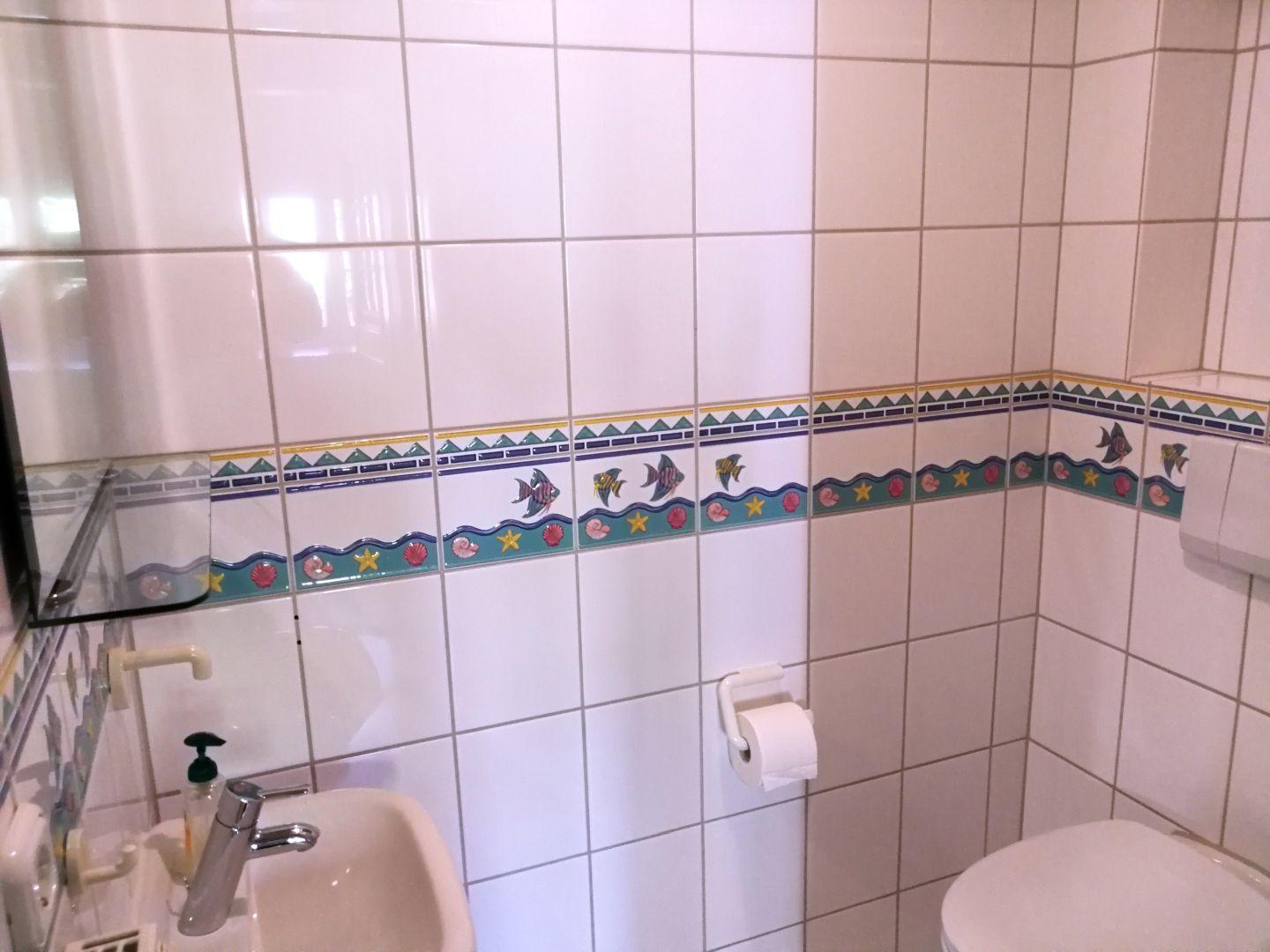 Badezimmer mit Fliesen, Waschbecken, Toilette und Toilettenpapierhalter.