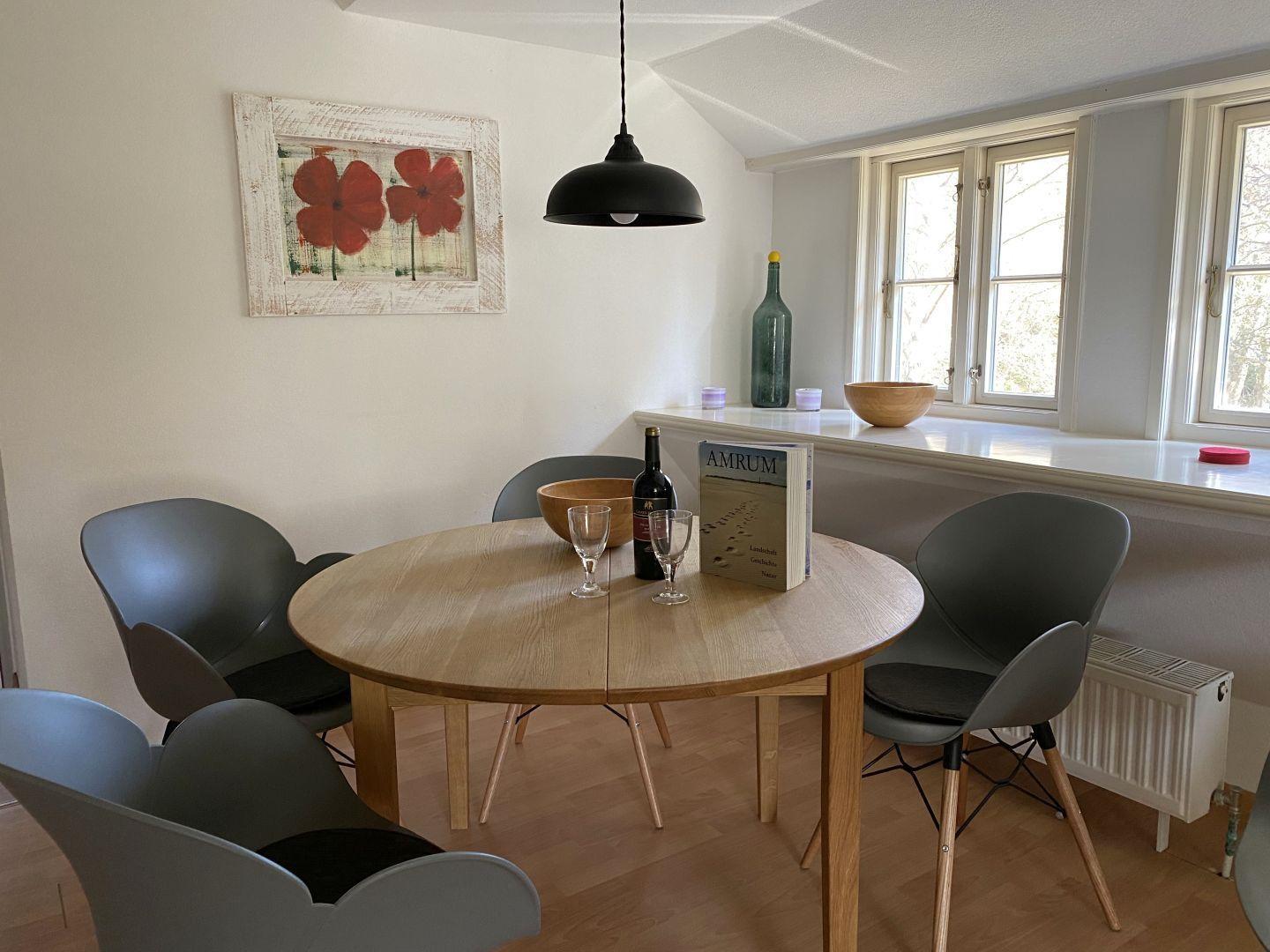 Runder Holztisch mit Stühlen, Fensterbank und Wandbild mit roten Blumen.