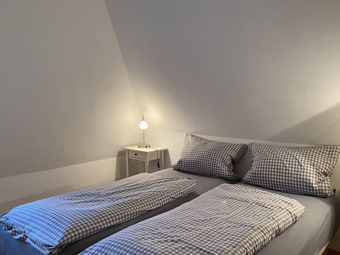 Doppelbett mit kariertem Bettwäsche und Nachttisch mit Lampe unter schräger Decke.