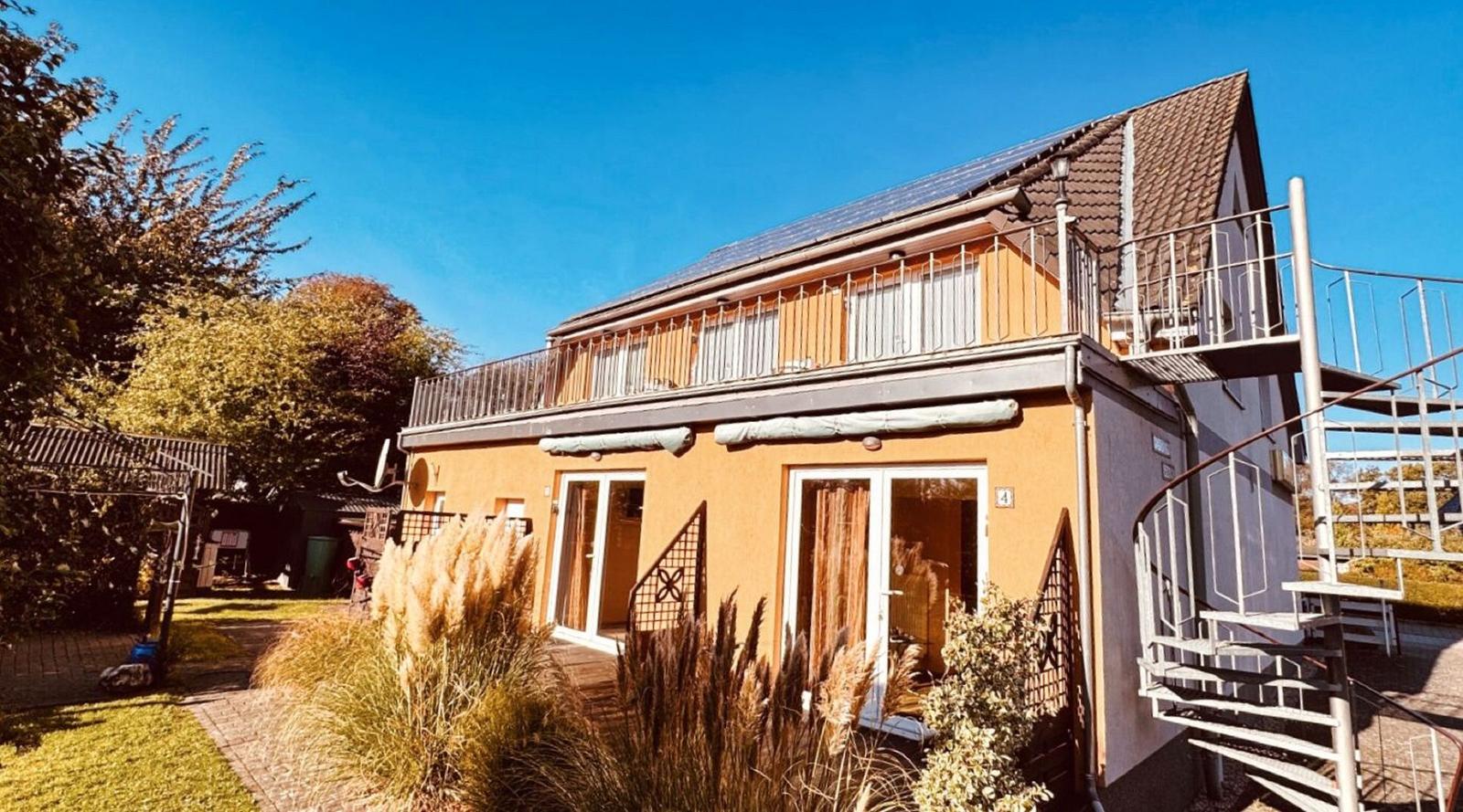 Haus mit Balkon und螺旋楼梯, umgeben von Grünflächen und Bäumen unter blauem Himmel.