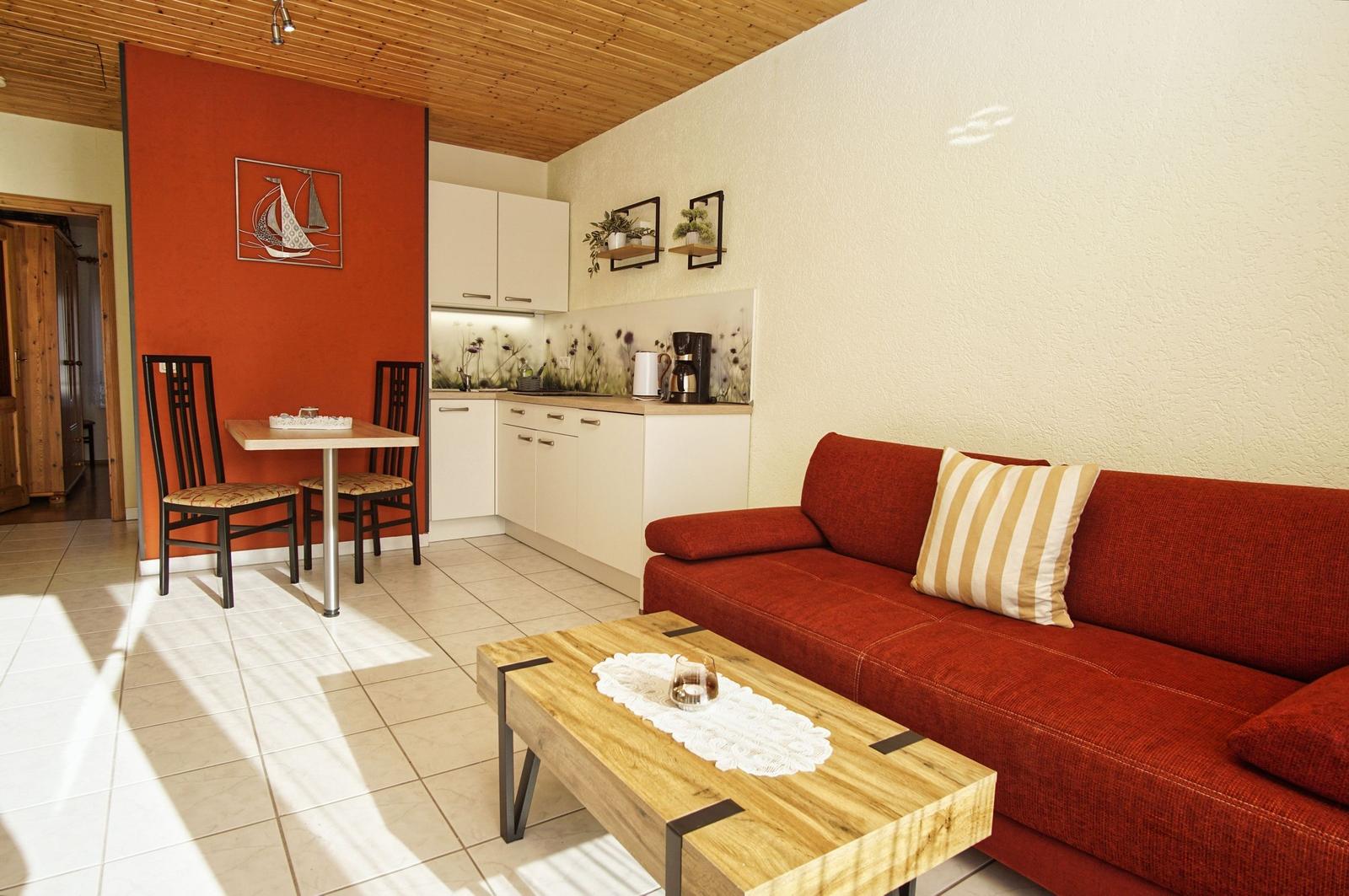 Wohn- und Küchenbereich mit rotem Sofa, Holztisch und weißen Schränken.