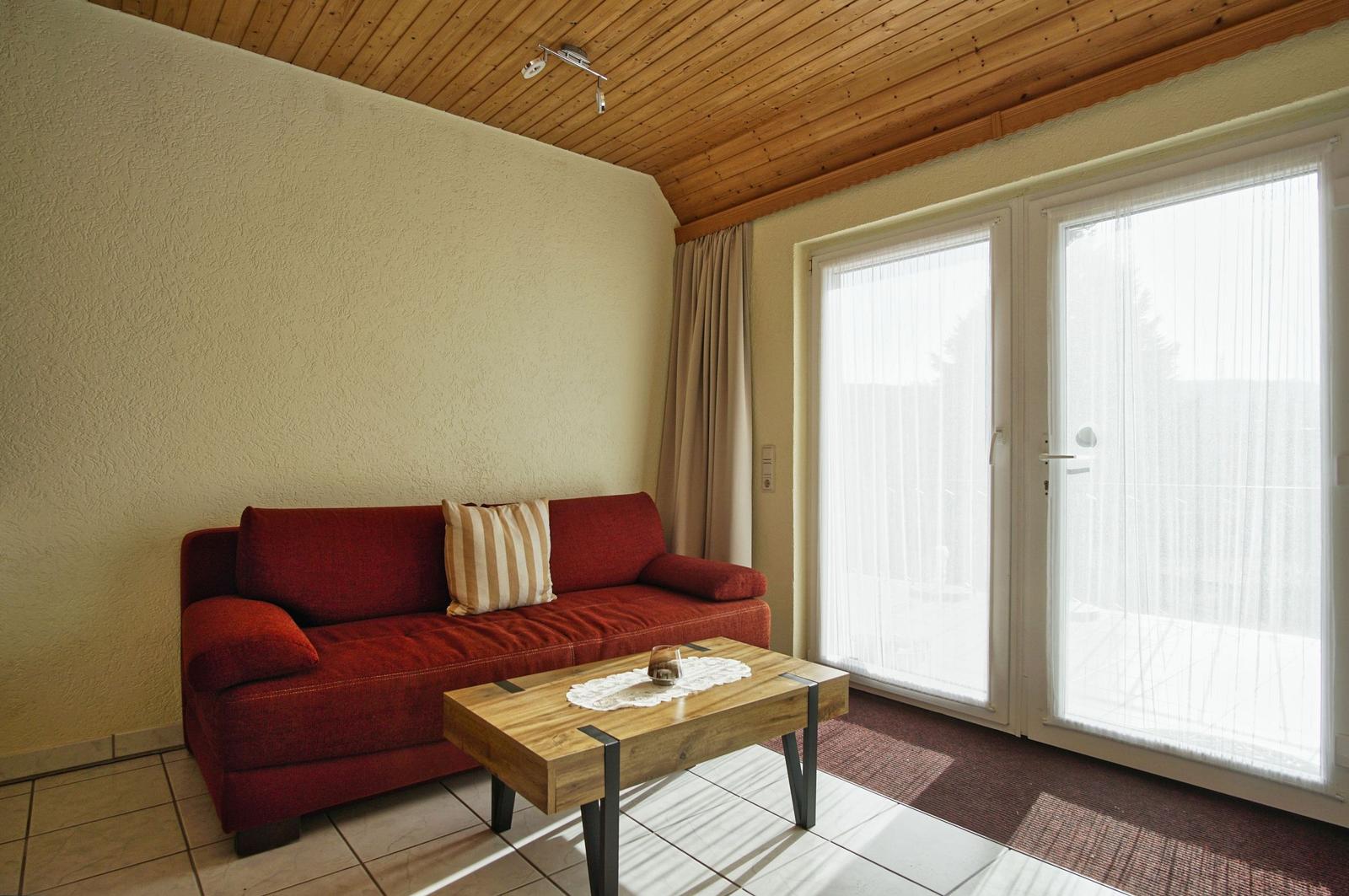 Ein Wohnzimmer mit rotem Sofa, Holztisch und Fenster zur Terrasse.