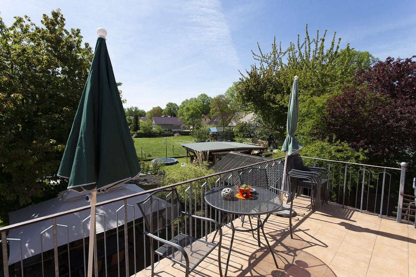 Terrasse mit Tisch, Stühlen und grünen Sonnenschirmen. Blick auf Garten und Bäume.