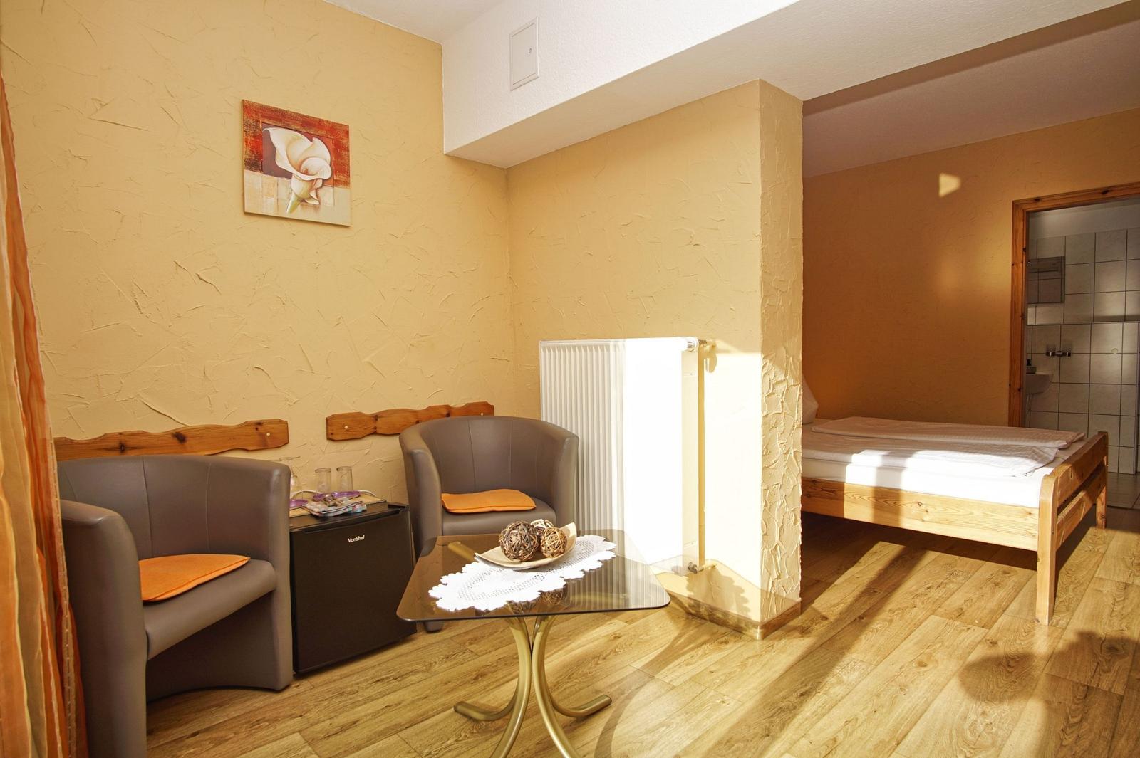 Zimmer mit Schlafbereich, zwei Sesseln, Tisch und Kühlschrank.