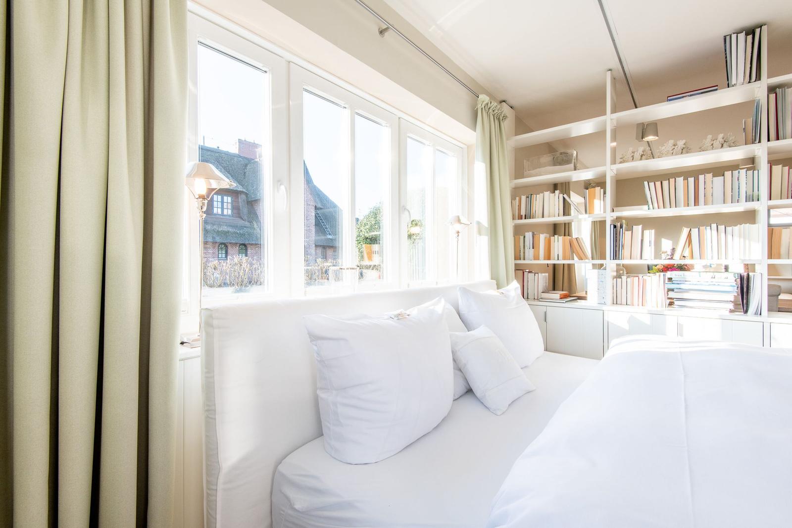Schlafzimmer mit weißem Bett, Bücherregal und Fensterblick auf Dachhäuser.