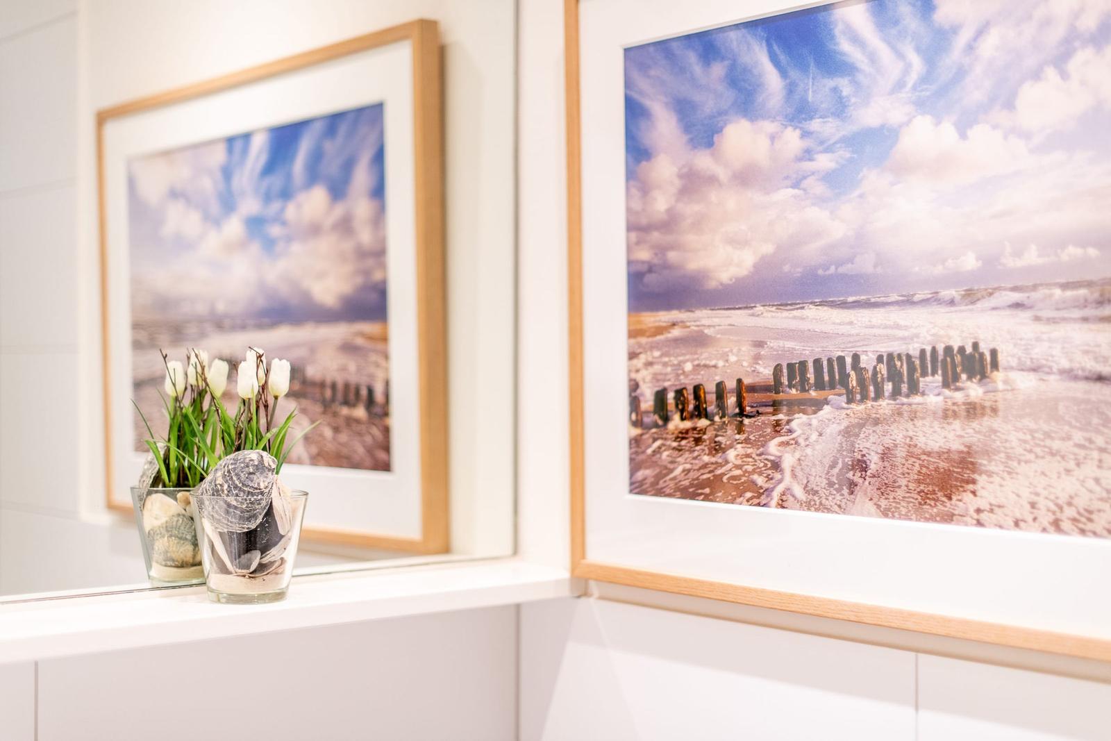 Zwei gerahmte Strandbilder hängen an der Wand. Davor steht eine Vase mit Tulpen und Muscheln.