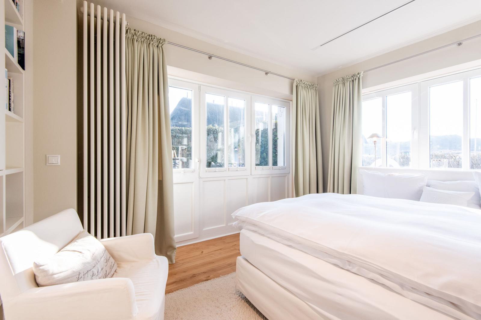 Schlafzimmer mit weißem Bett, Sessel und großen Fenstern.