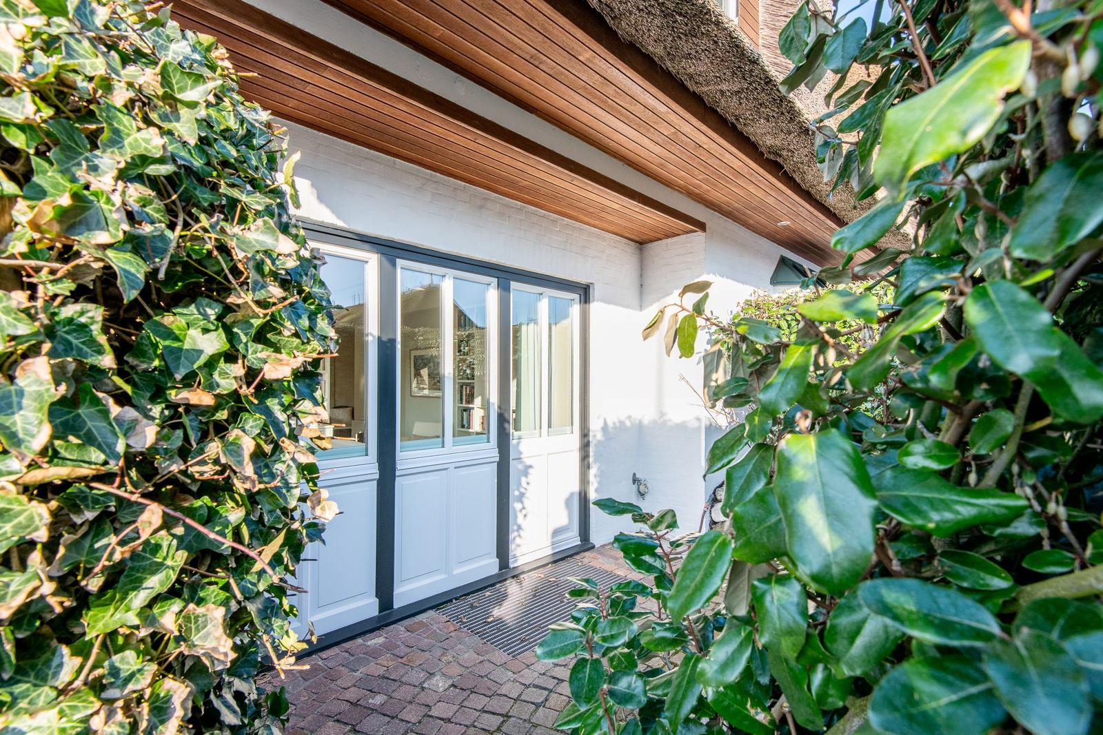 Haus mit weißen Türen und strohgedecktem Dach, umgeben von grüner Vegetation.