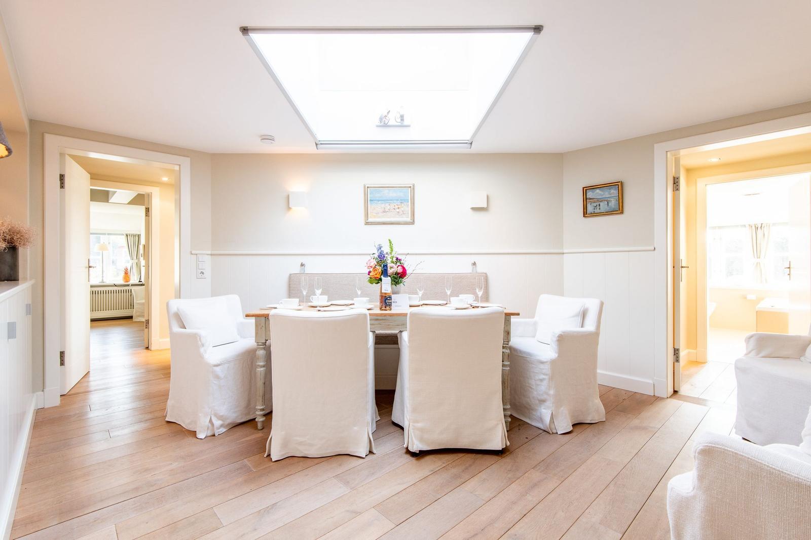 Esszimmer mit Tisch, Stühlen und Deckenfenster. Holzboden und Wandbilder sichtbar.