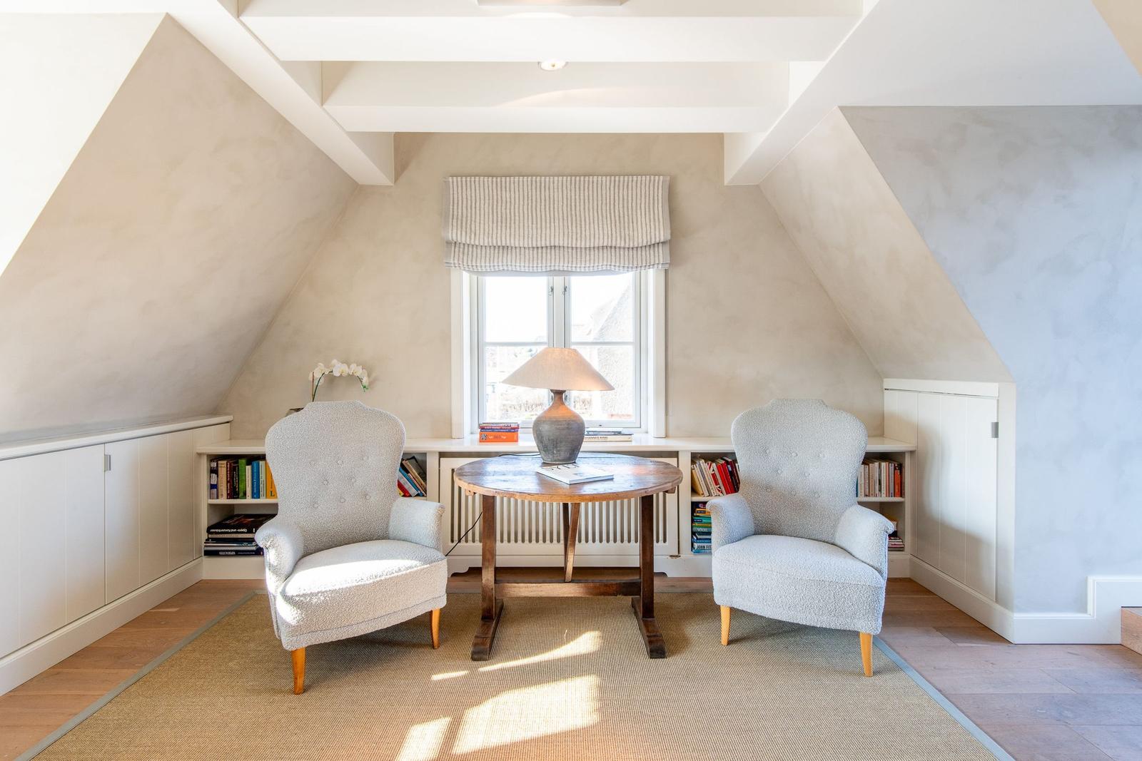 Zwei Sessel und ein Tisch stehen vor einem Fenster in einem Dachzimmer mit Bücherregalen.