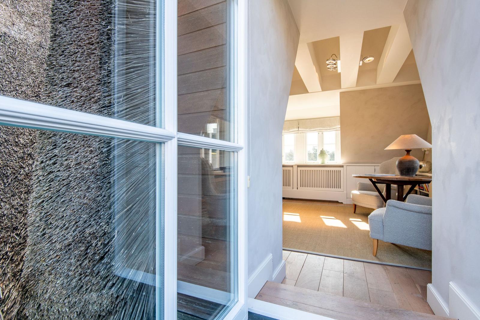 Durch einen Flur mit Holzboden und weißen Wänden gelangt man in ein Wohnzimmer mit Sitzgelegenheiten.