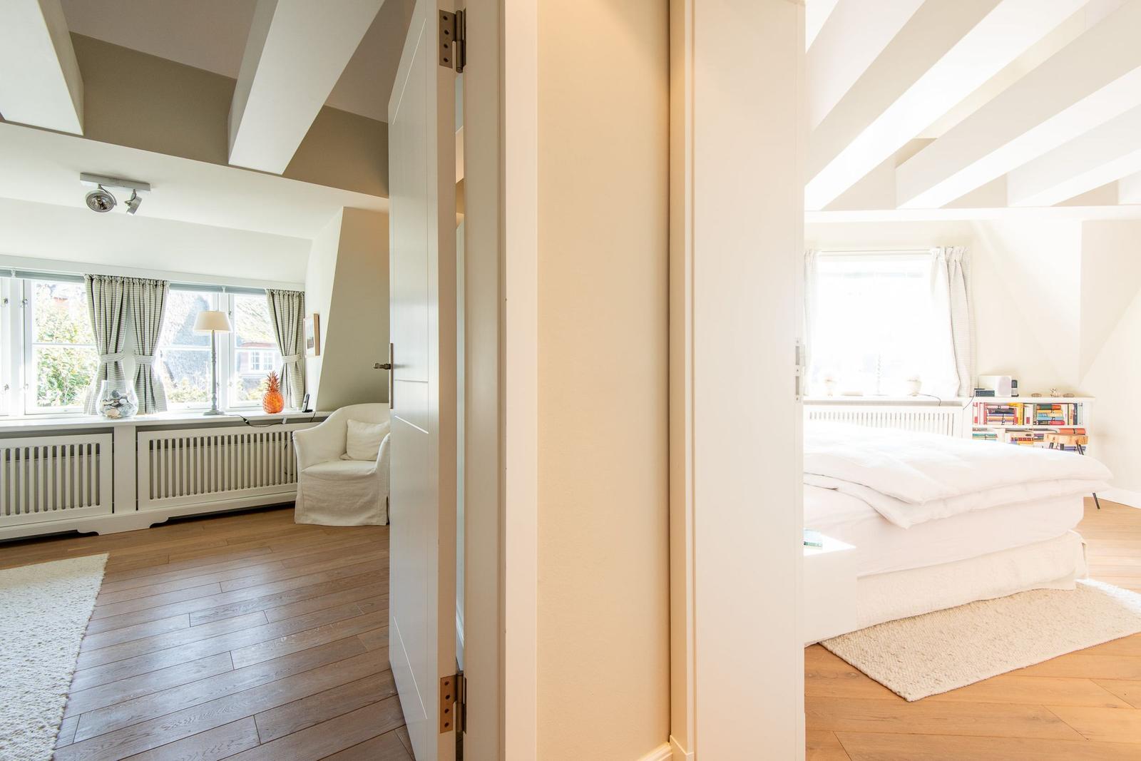 Zimmer mit Bett und Bücherregal. Durchgang zu Wohnbereich mit Fenster und Sessel.