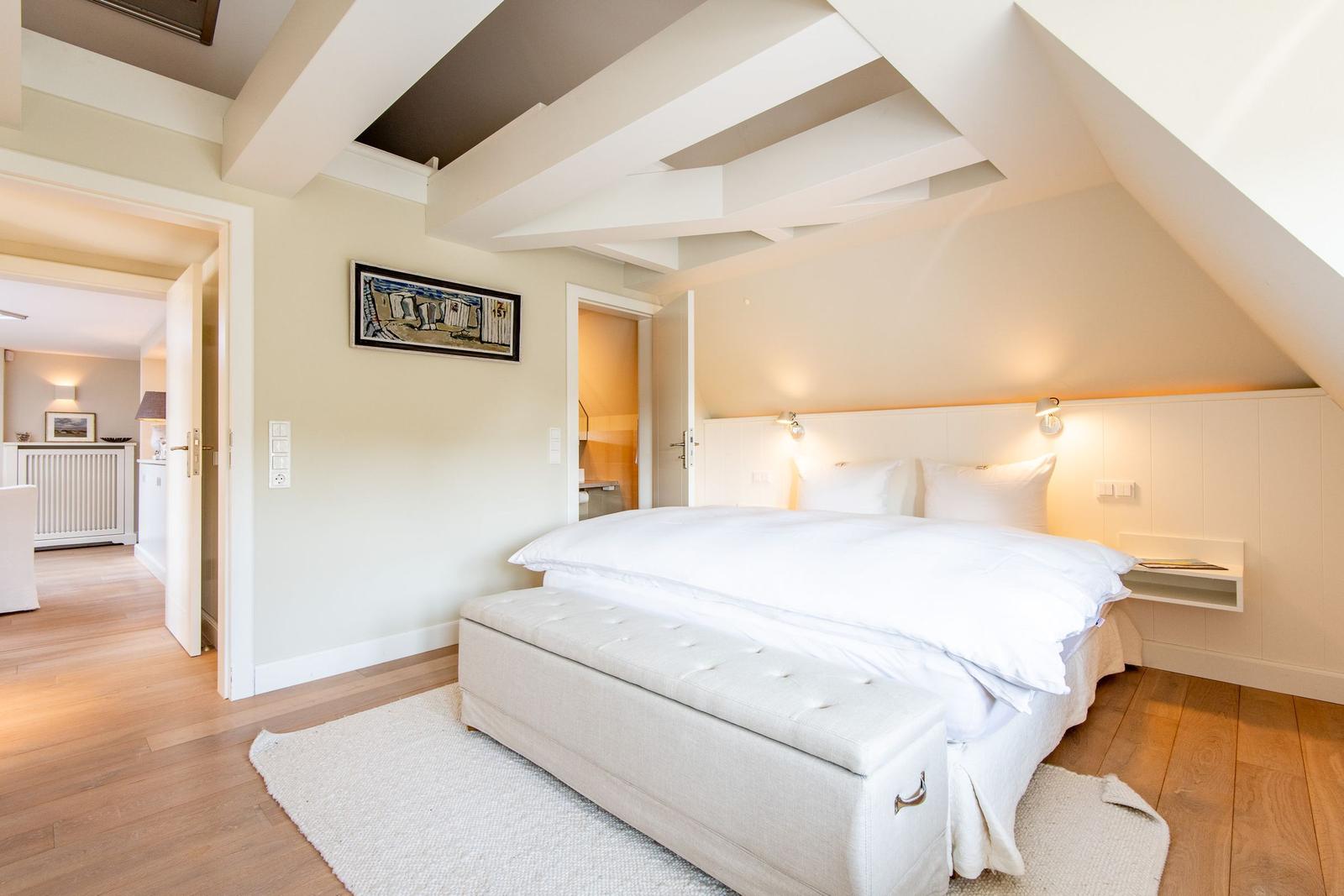 Schlafzimmer mit Doppelbett, Holzboden und weißen Wänden. Deckenbalken und Fenster sichtbar.