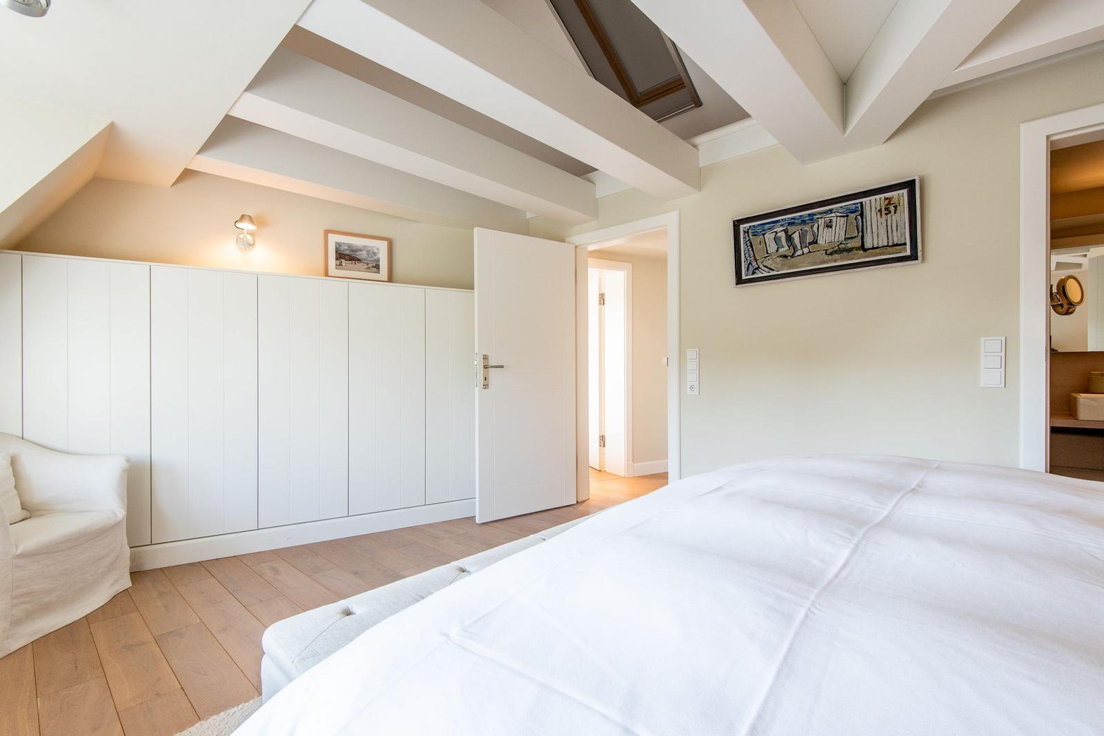 Schlafzimmer mit weißem Bett, Holzboden und Deckenbalken. An der Wand hängt ein Bild.