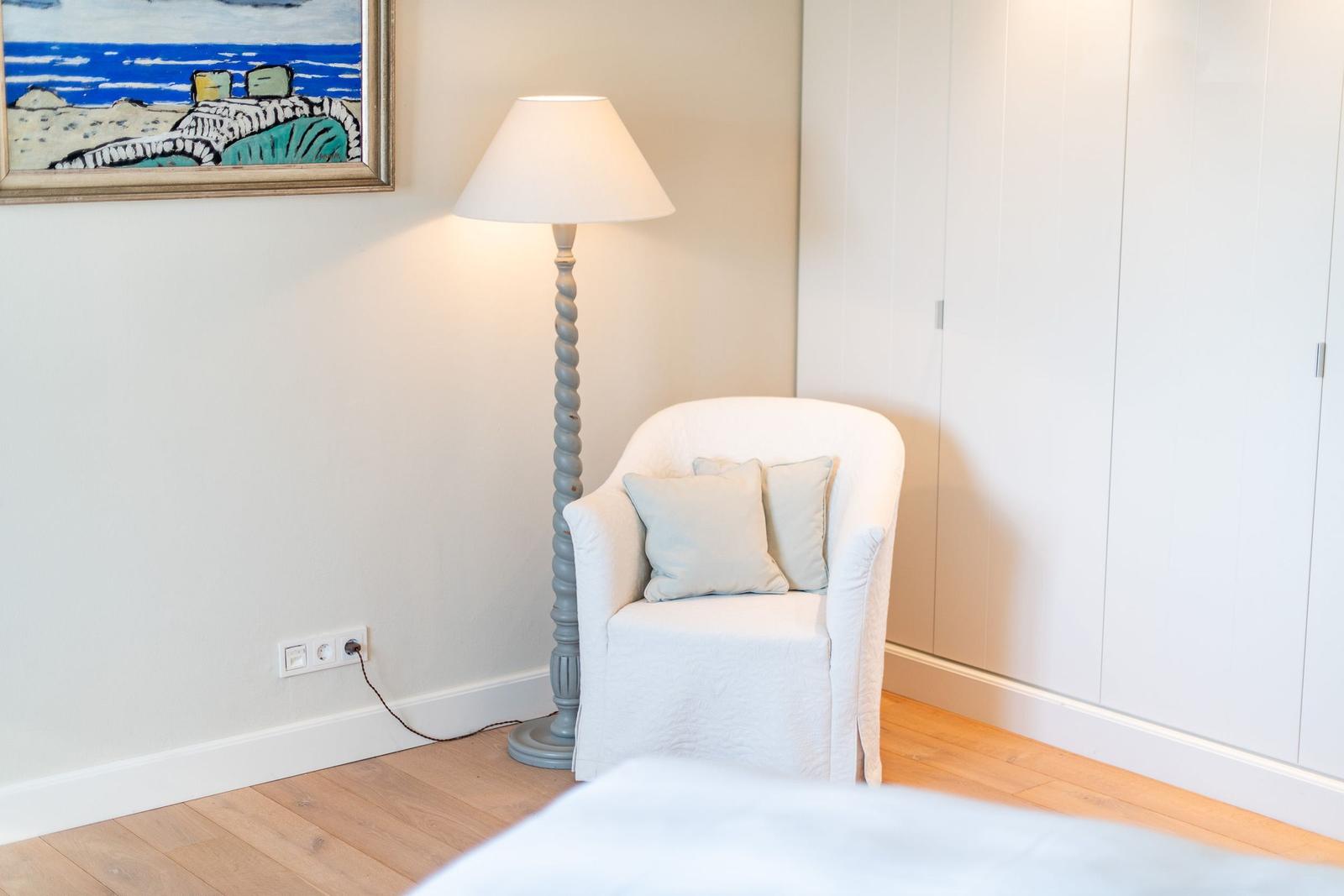 Ein weißer Sessel mit Kissen steht neben einer Stehlampe und einem Schrank.