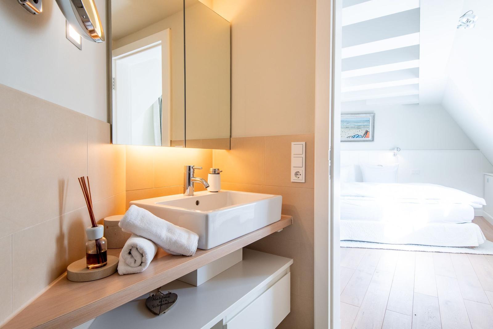 Badezimmer mit Waschbecken und Blick in Schlafzimmer mit Dachschrägen.