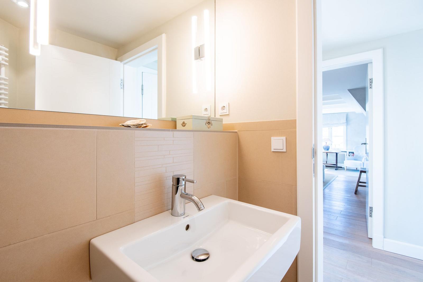 Bad mit Waschbecken, Spiegel und Lichtleisten. Tür führt in Wohnbereich.