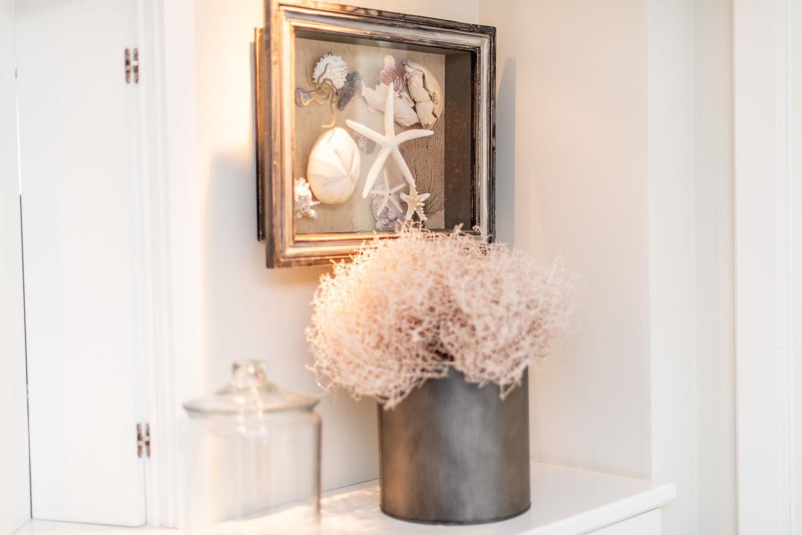 Wandbild mit Meereskunst, Vase mit Trockenpflanze und Glasbehälter auf weißem Regal.
